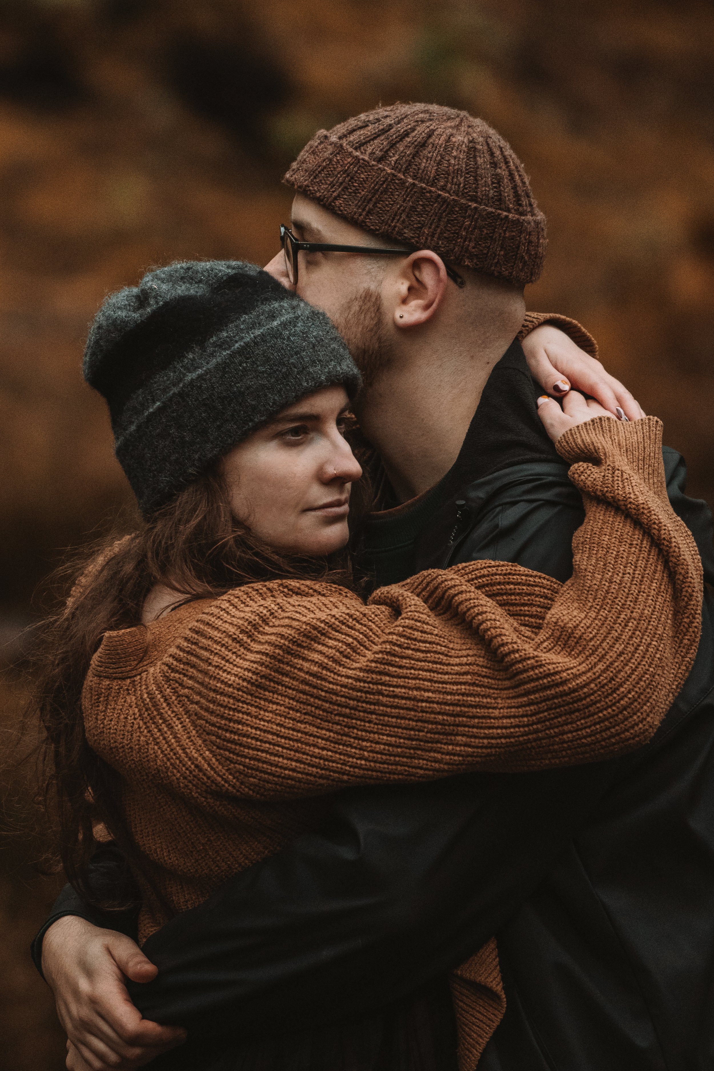 A couple embracing outdoors, the woman with long brown hair and a gray beanie, and the man with glasses, a beard, and a brown knit cap, both wearing warm brown sweaters.