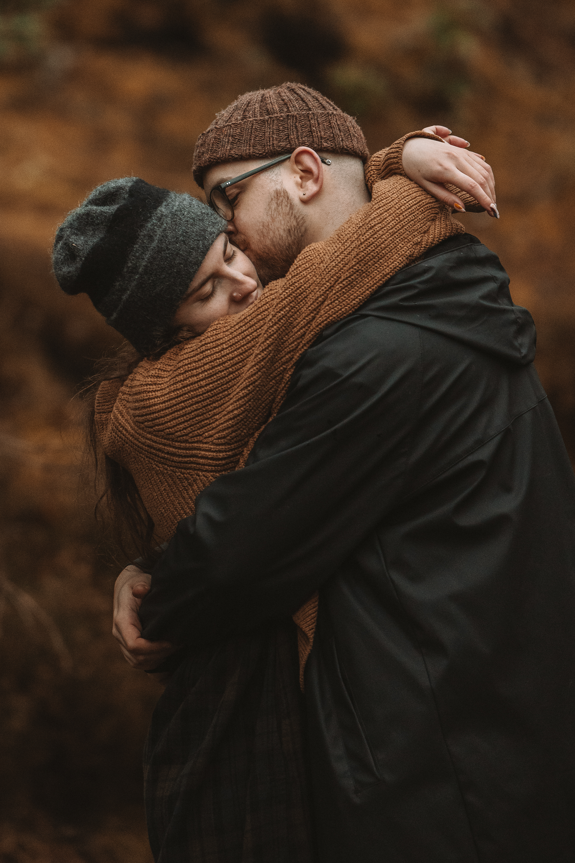A young couple embracing outdoors during fall, with the man kissing the woman and both wearing warm clothing and hats.