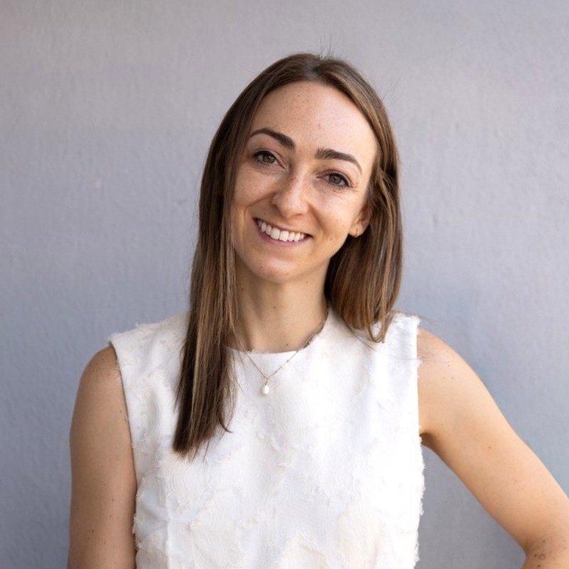 A young woman with long brown hair smiling at the camera against a plain gray wall.