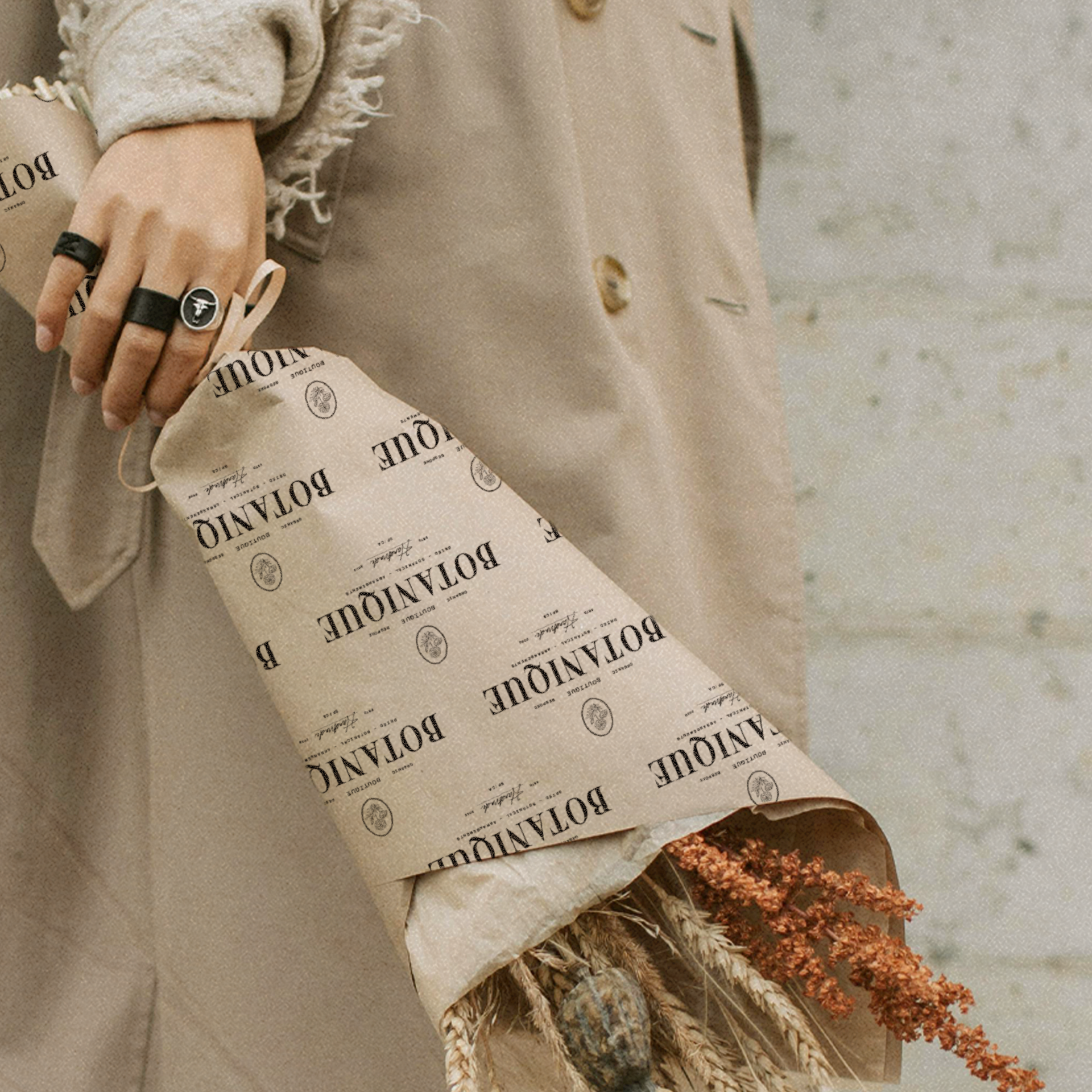 A person holding a bouquet of dried flowers wrapped in patterned paper, with a background of a beige coat and a grey wall.