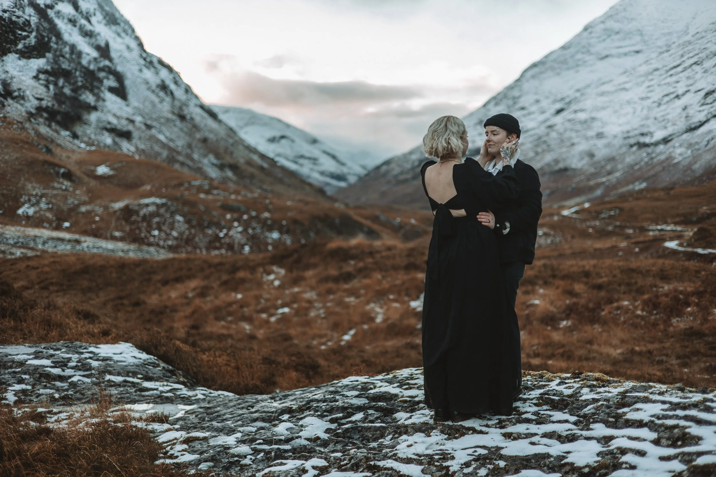 Two women standing close together embracing in a mountain valley with snow-covered peaks, brown grasses, and rocky terrain in the background during daylight.