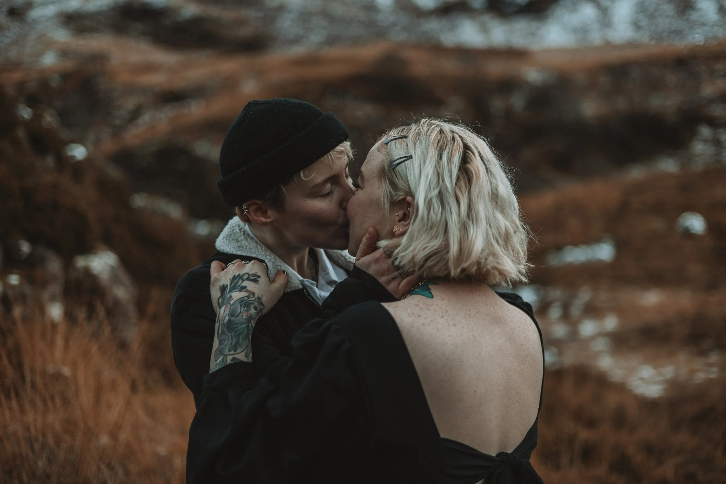 A couple sharing a kiss outdoors, with the woman having blonde hair, tattoos on her arm, and gratified expression, and the man wearing a black beanie, earrings, and a black hoodie, surrounded by brownish, blurry natural background.