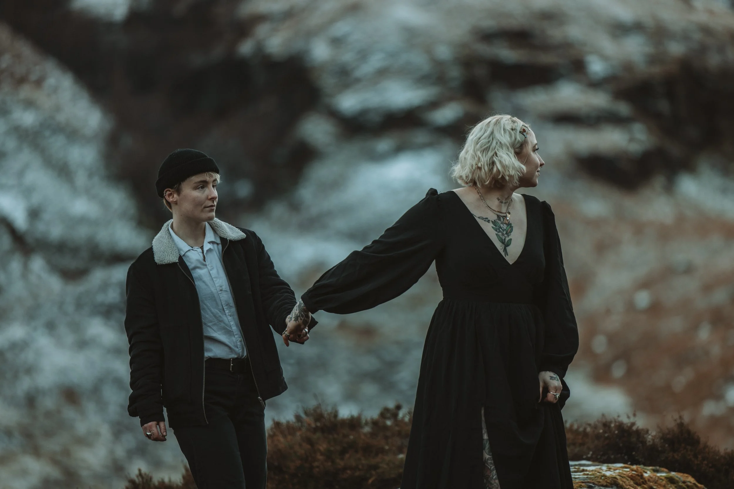 Two women walking outdoors during dusk, holding hands, with a blurred rocky background. One woman has short dark hair, wears a beanie, a black jacket, and a white shirt. The other woman has shoulder-length platinum blonde hair, tattoos on her chest a