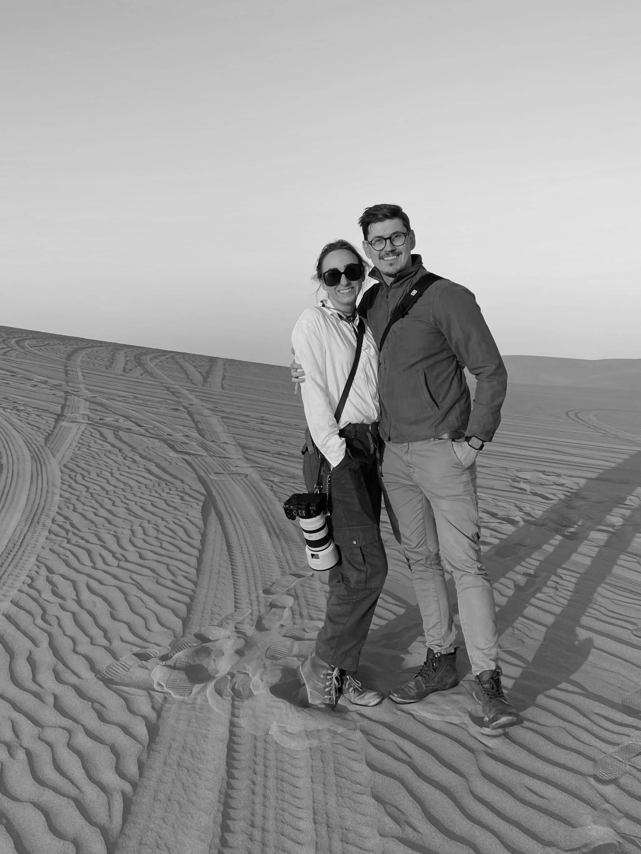 Black and white photo of a smiling man and woman standing close together in a desert landscape with tire tracks in the sand. The man has short dark hair, glasses, and is wearing a jacket and khakis. The woman has her hair tied back, sunglasses, and is wearing a light jacket, with a camera hanging around her neck.