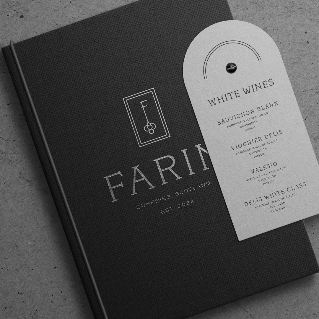 Black hardcover menu with the word "FARING" and the text "DUMFRIES, SCOTLAND, EST. 2024" next to a white wine list on a grey surface.