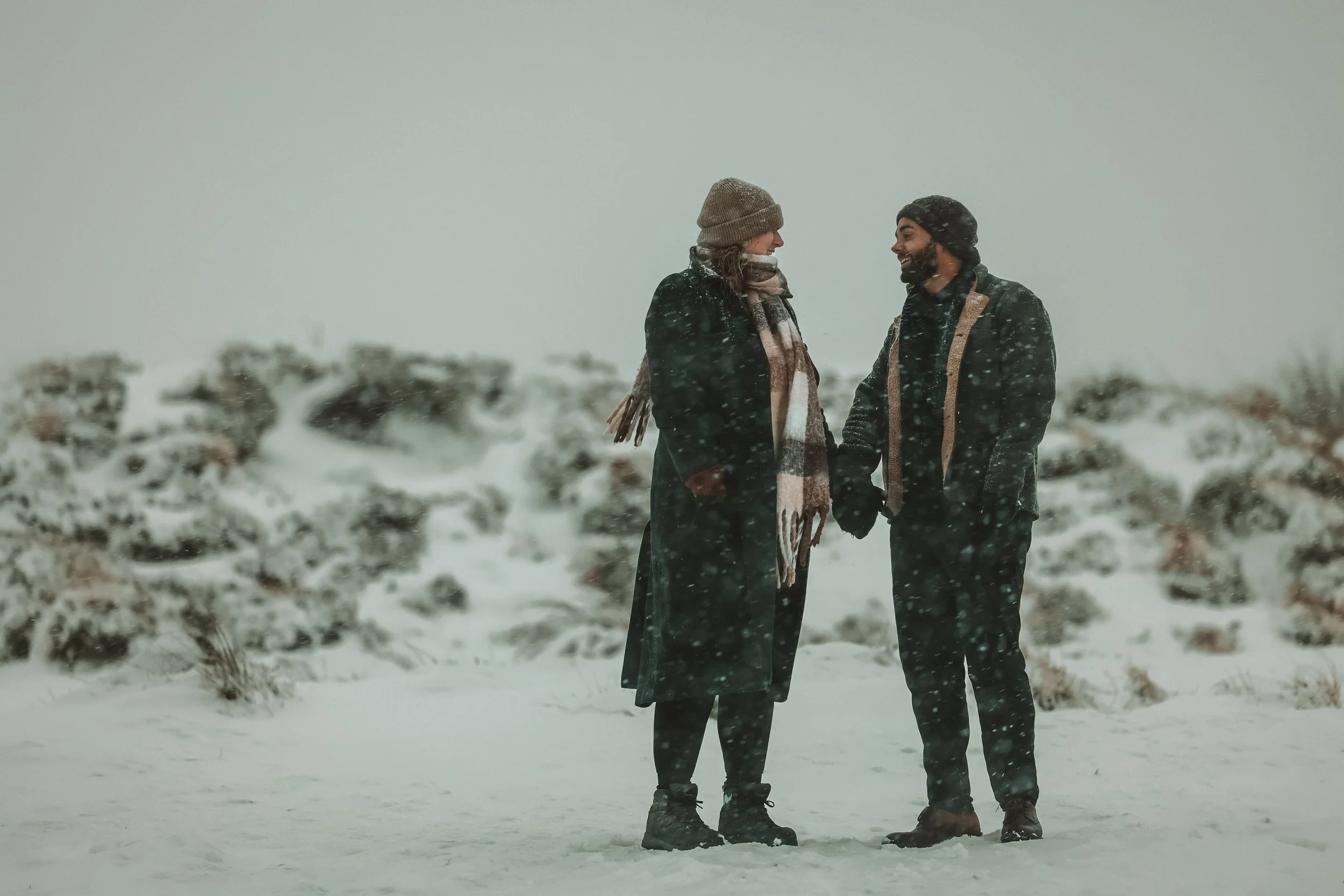A couple standing close together outdoors in a snowy landscape, smiling and holding hands, dressed in warm winter clothing including hats and coats, with snow falling around them.