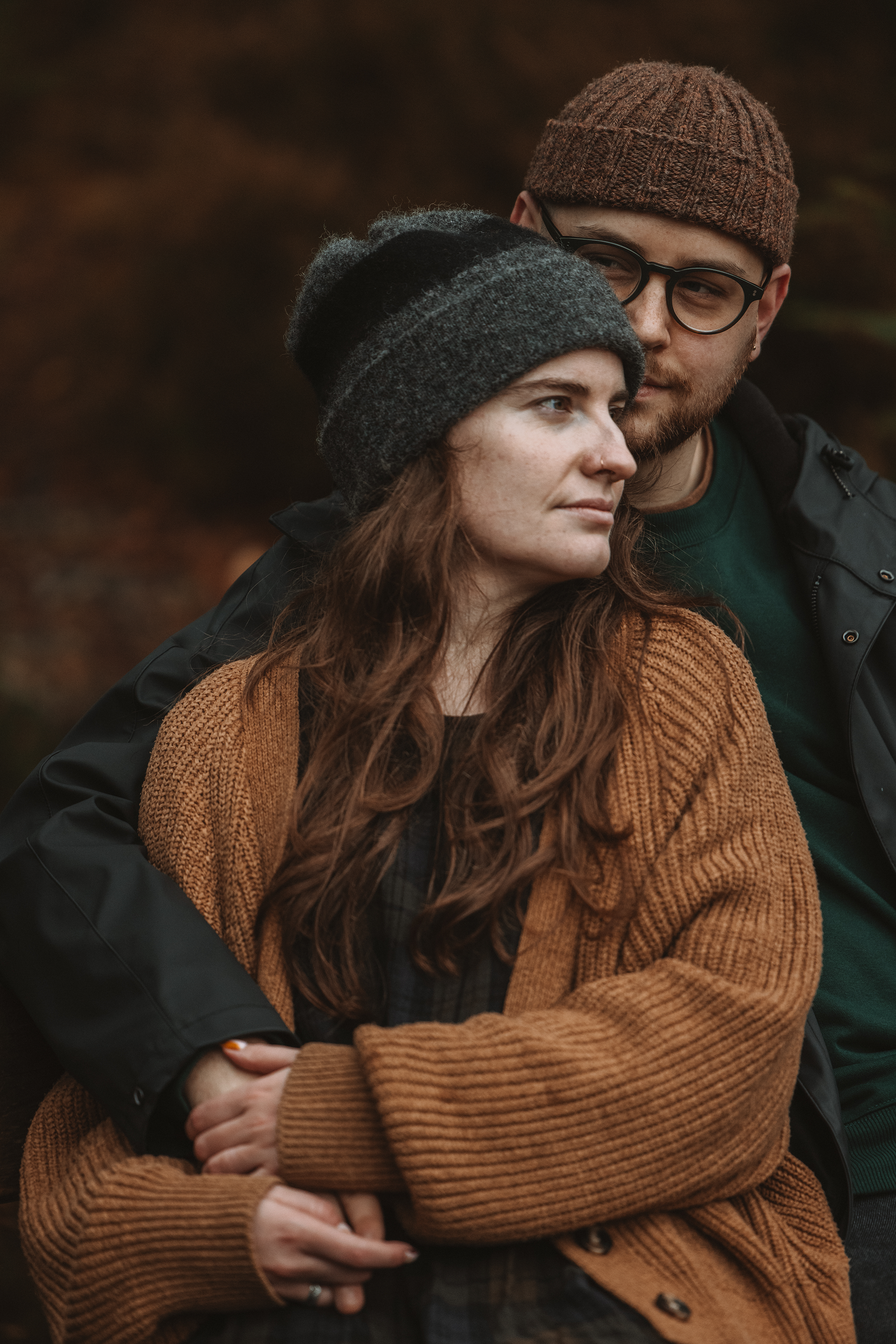 A young woman with long reddish hair, wearing a dark gray beanie and a brown sweater, stands with crossed arms. A young man with short hair, glasses, a brown knit cap, and a black jacket stands behind her, looking at her.