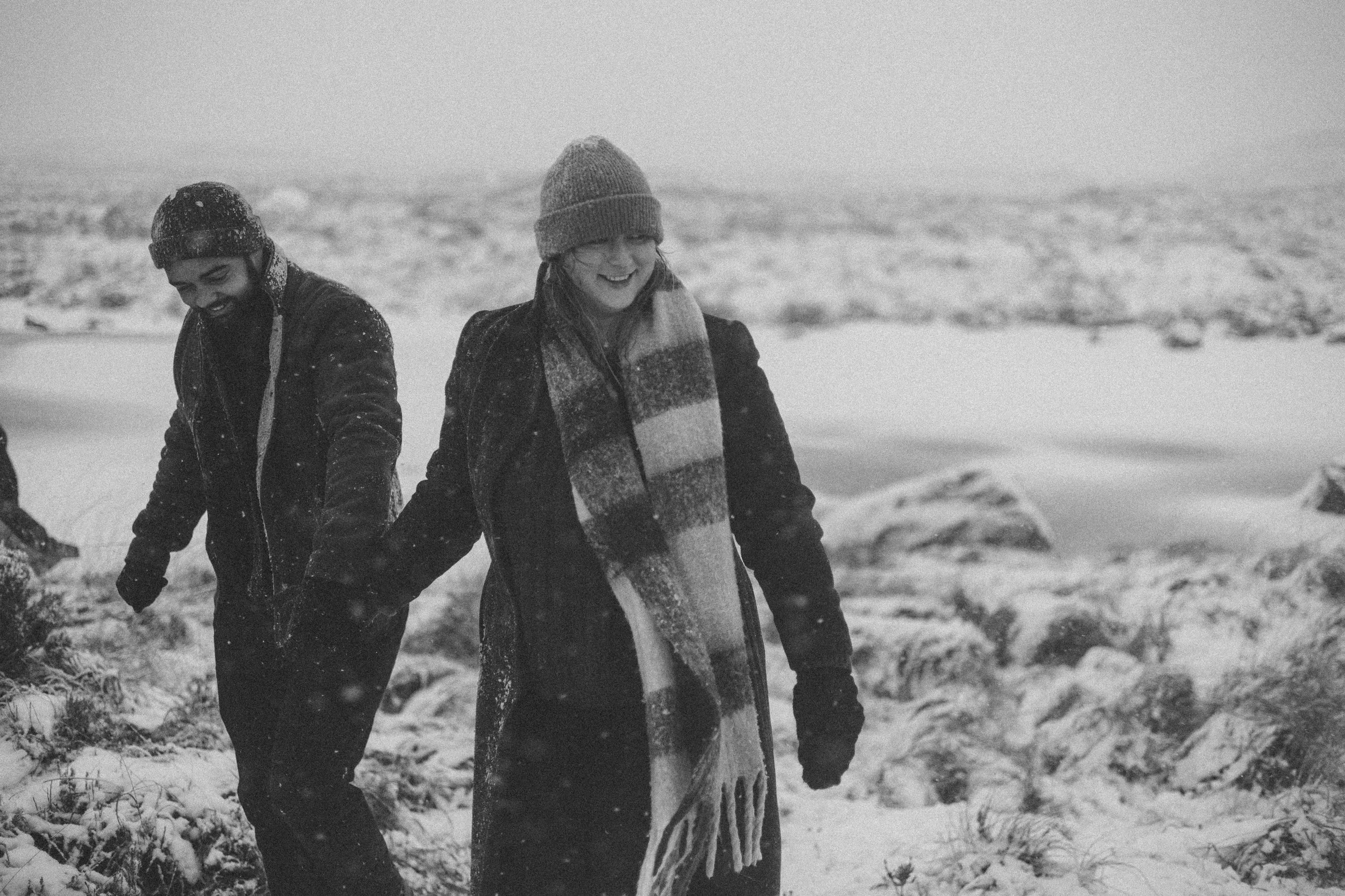 Two smiling people, one man and one woman, walking in a snowy outdoor landscape. The person on the right is holding a large, striped surfboard. Both are dressed warmly with hats and jackets.