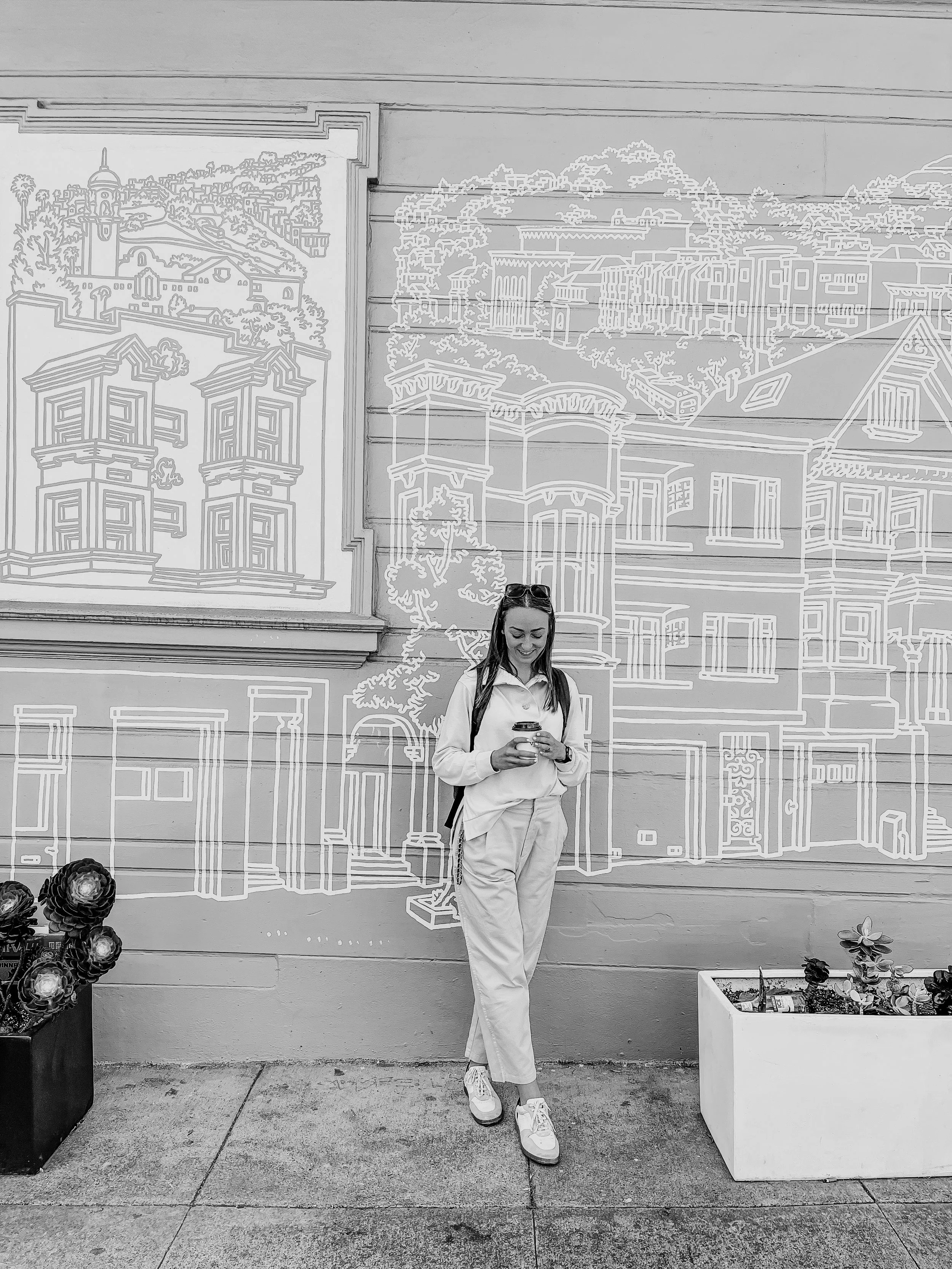 A woman with long dark hair, wearing a light-colored sports jacket and pants, leaning against a wall with street art outlines of buildings and trees, smiling and looking at her phone. She is standing on a sidewalk with plants, and there are decorative objects on the ground, with a pair of sunglasses on her head.