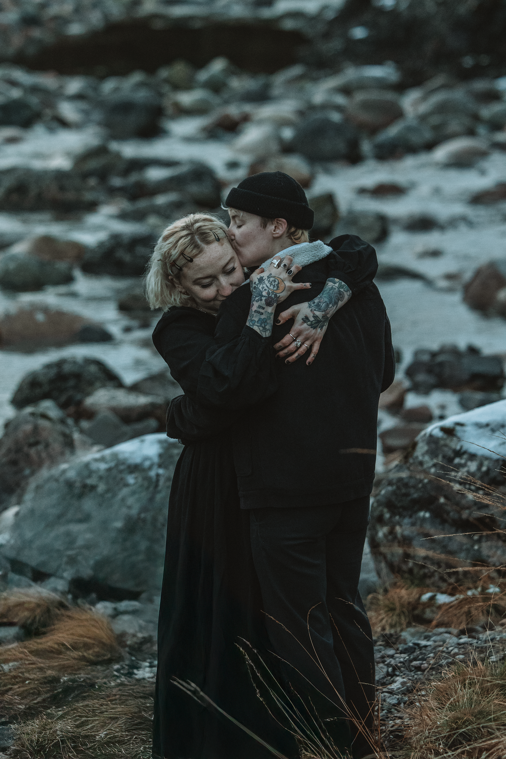 A couple hugging and kissing in a rocky outdoor setting, with the man kissing the woman's forehead as she smiles.