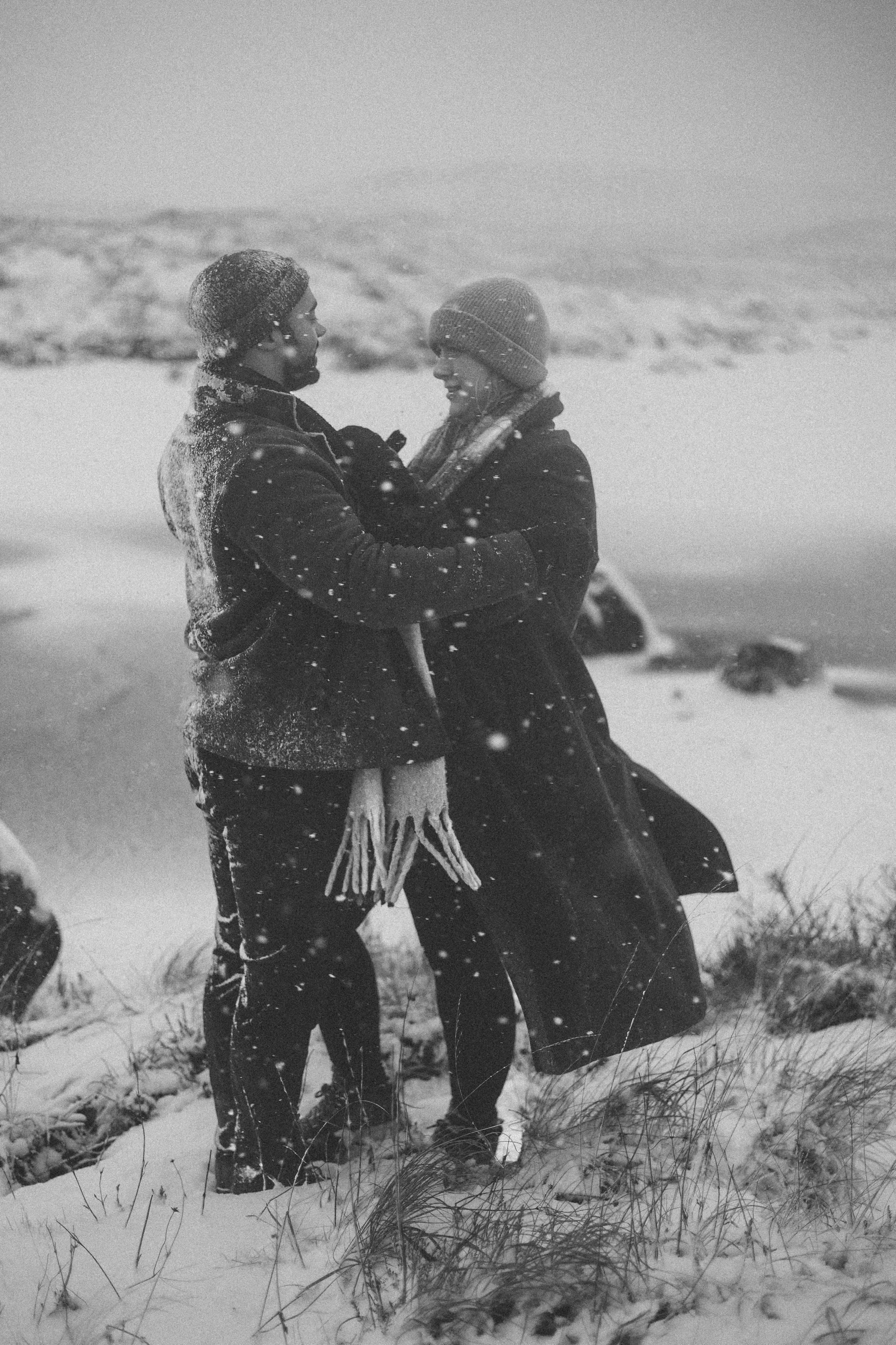 A black and white photograph of two people standing close in a snowy outdoor setting, smiling and looking at each other, with snow-covered ground and distant trees in the background.