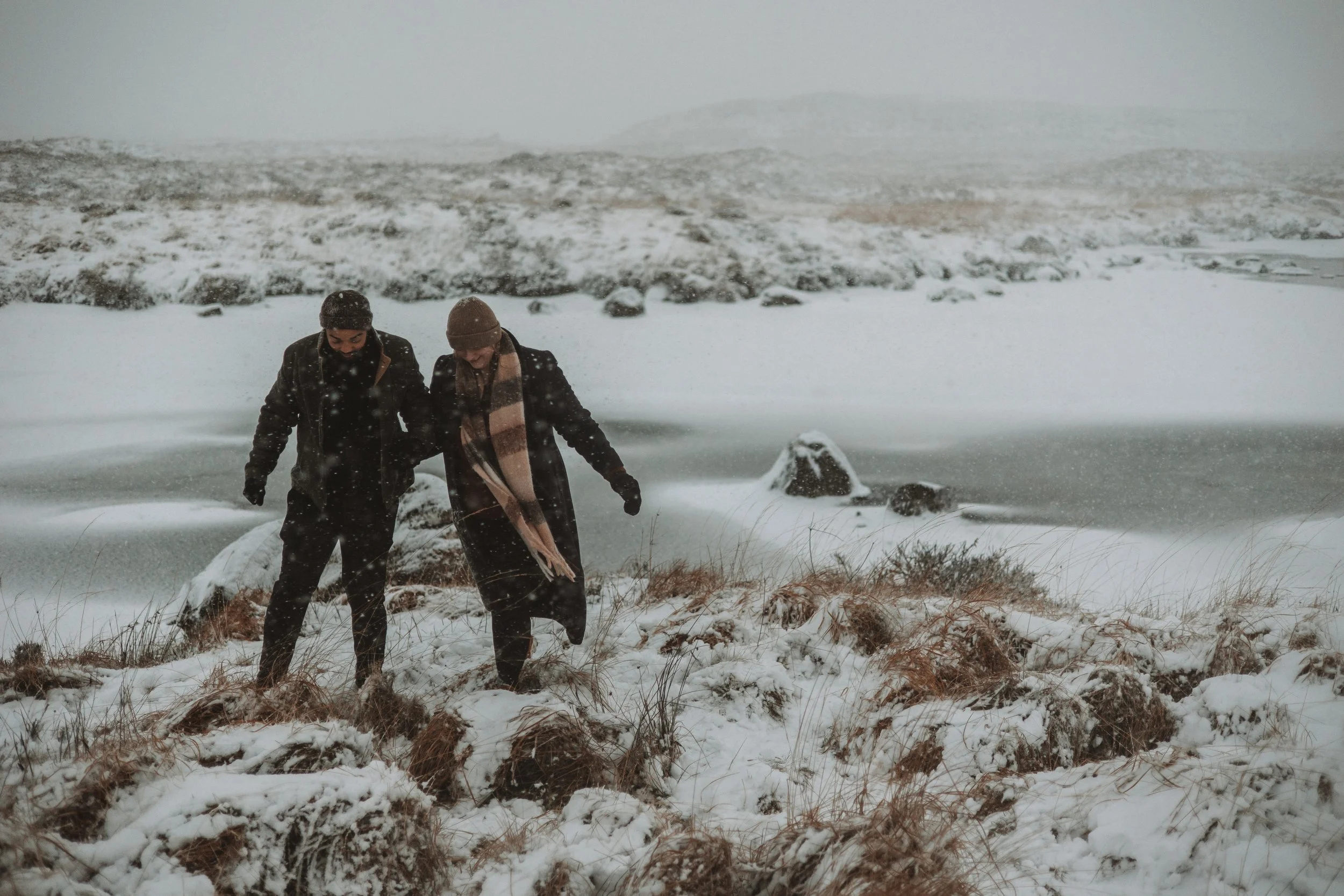 Two people dressed in winter clothing walking through snow-covered ground near a frozen body of water during a snowstorm.