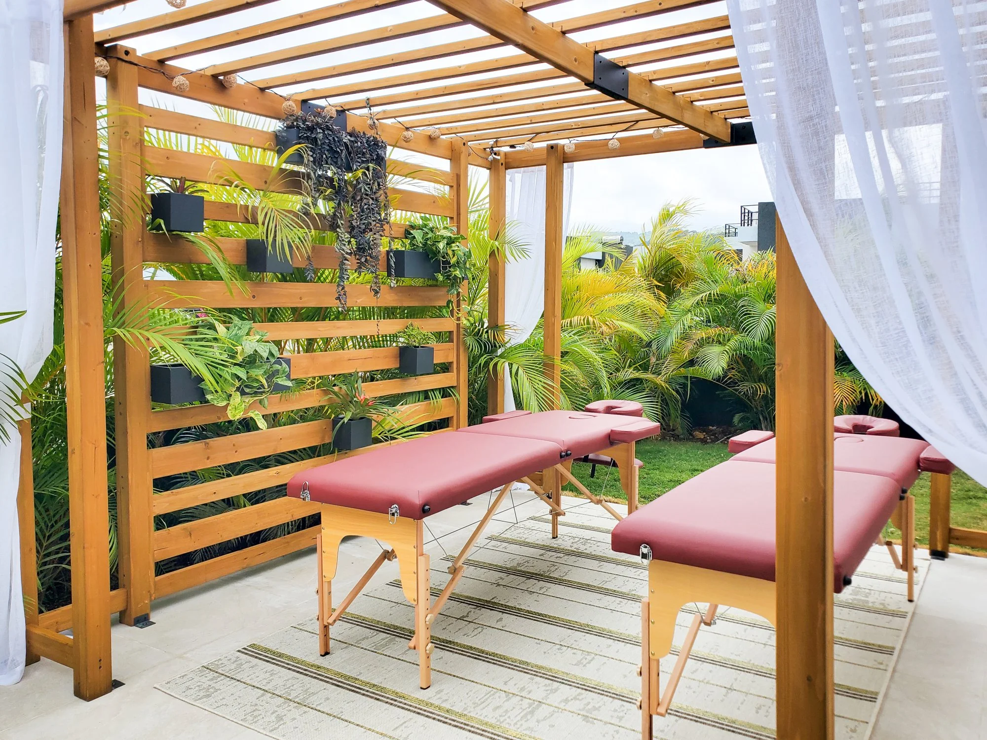 Two massage tables arranged beneath a wooden pergola with white curtains and lush tropical greenery for a private couples massage experience at Luxe Oasis villa in Ocho Rios, Jamaica.