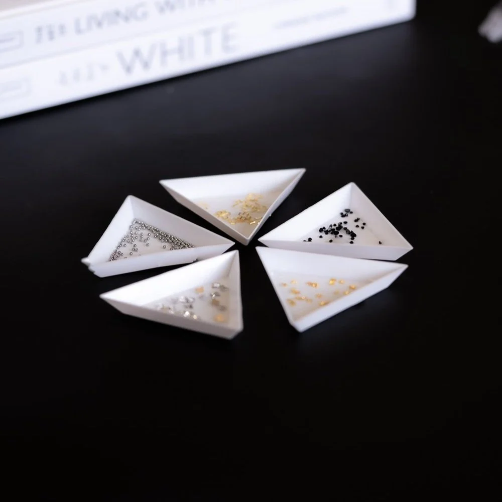 Five small white paper dishes with different types of nail art rhinestones and embellishments on a black surface.
