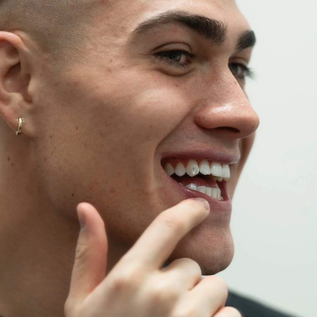Close-up of a smiling person with short hair, wearing a hoop earring, touching their lips with their fingers.