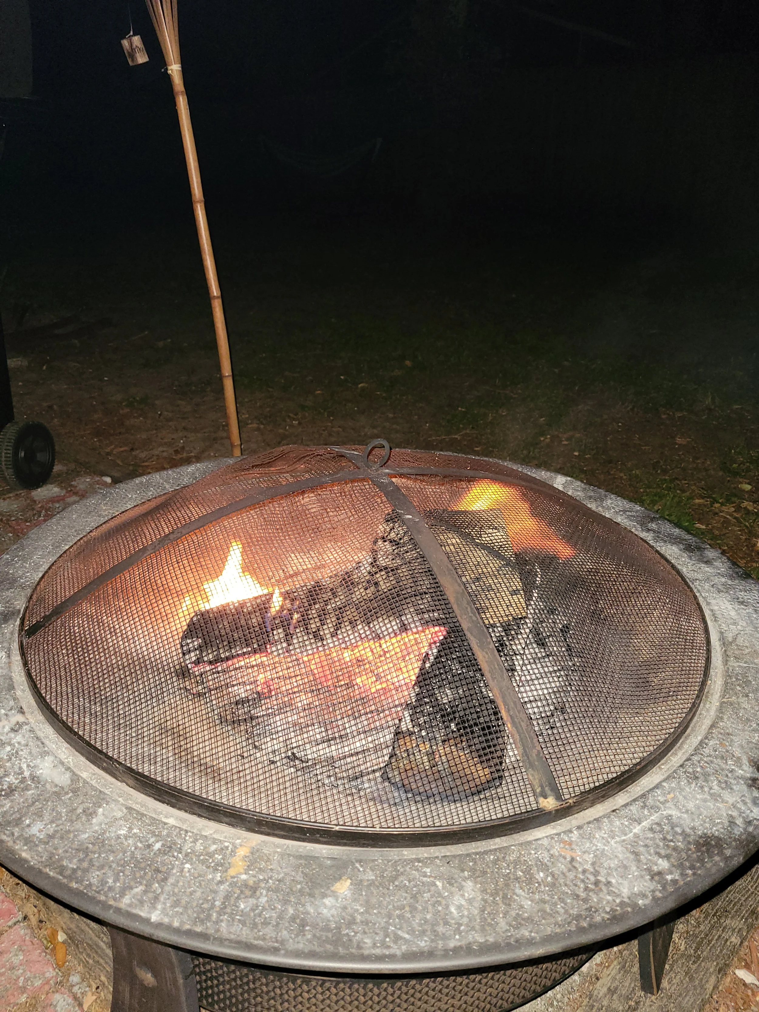 A metal fire pit with a mesh cover, containing burning logs and flames, in a dark outdoor setting.