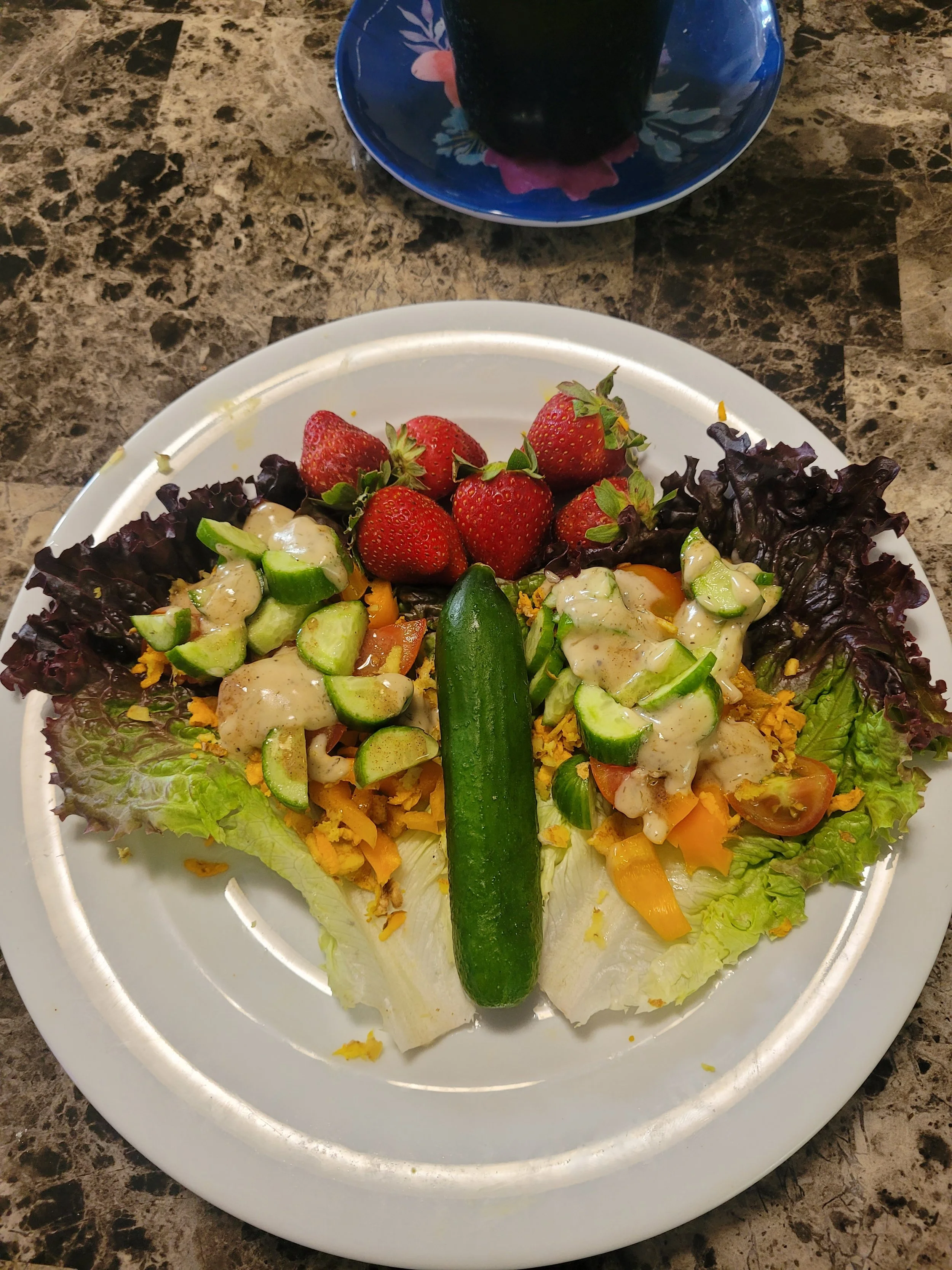Plate of fresh salad with lettuce, strawberries, sliced cucumbers, grated cheese, tomatoes, and dressing, topped with a whole cucumber.