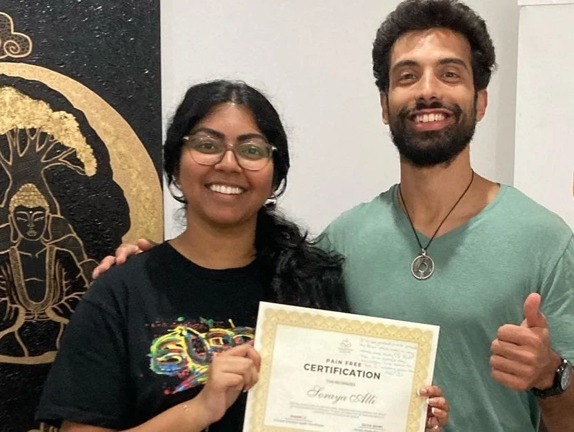 Two people smiling, with one holding a certificate that reads "Pain Free Certification." They are standing in front of a decorative wall with a Buddha motif.