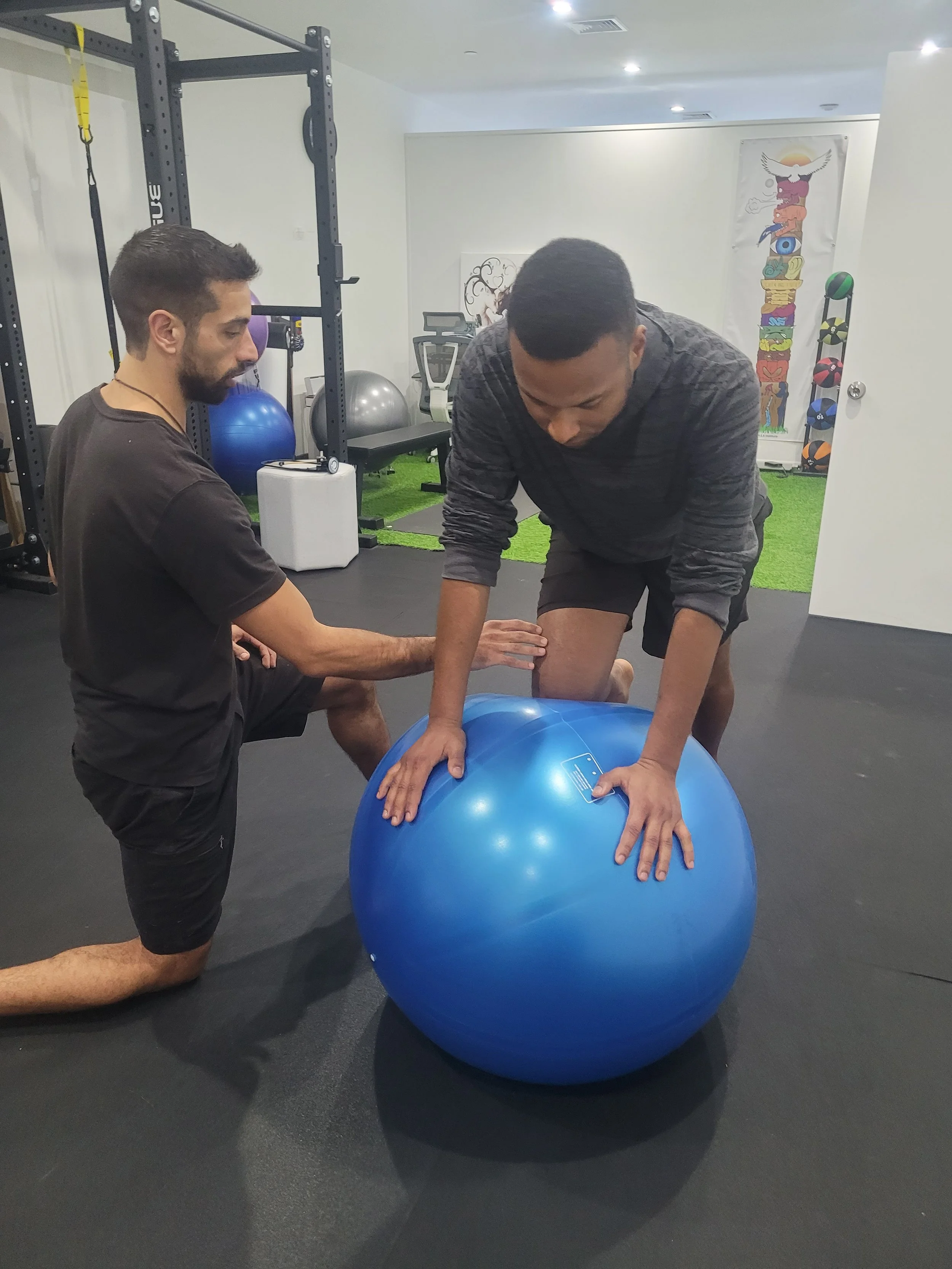 A physical therapist assists a man balancing on a blue exercise ball in a gym setting. The room contains fitness equipment such as a squat rack, exercise balls, and a stationary bike. The floor is made of rubber matting, and there is a decorative wall hanging in the background.