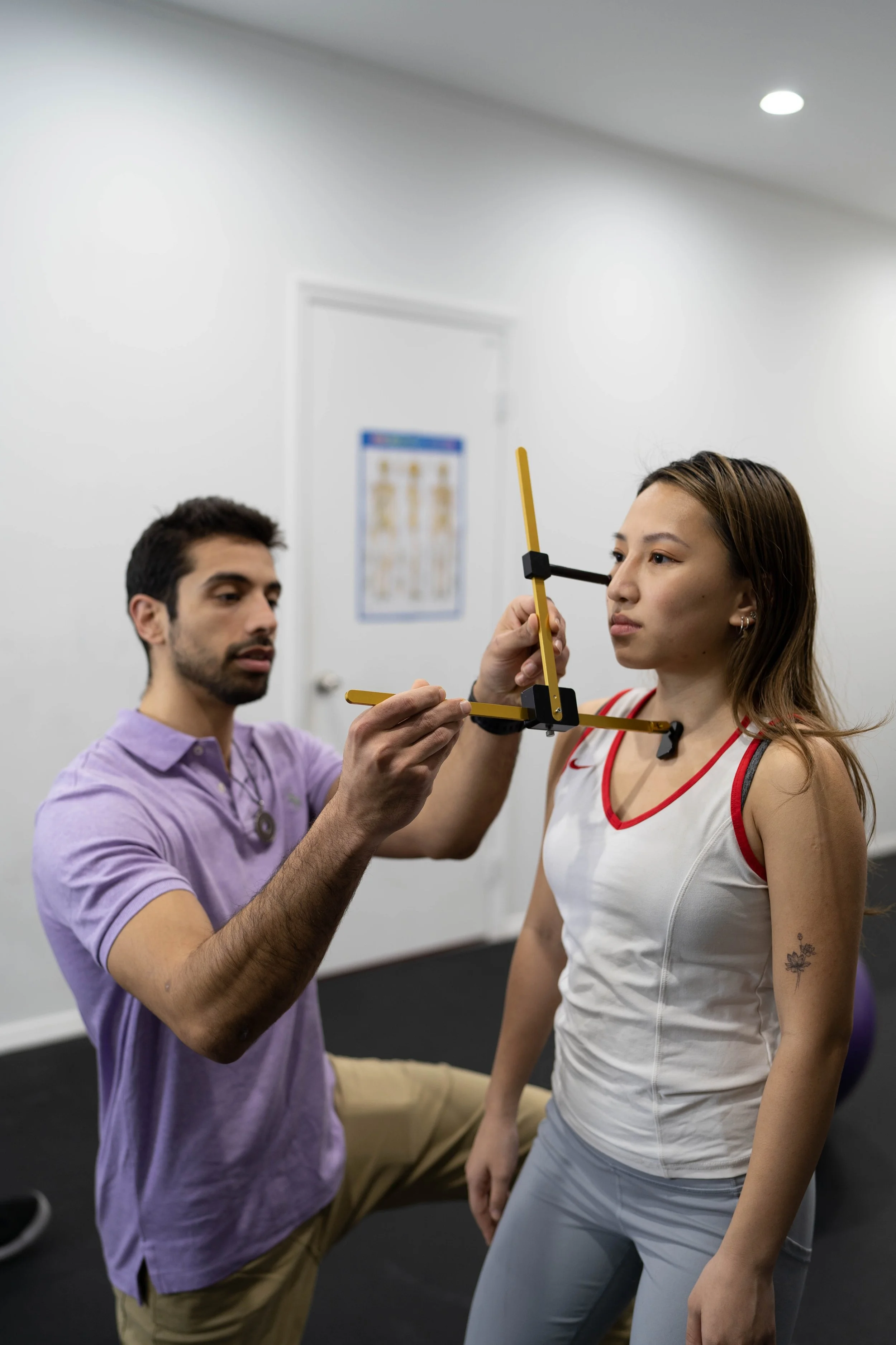 A man in a purple shirt using a measuring tool to assess a woman's posture or alignment in a gym or clinic setting.