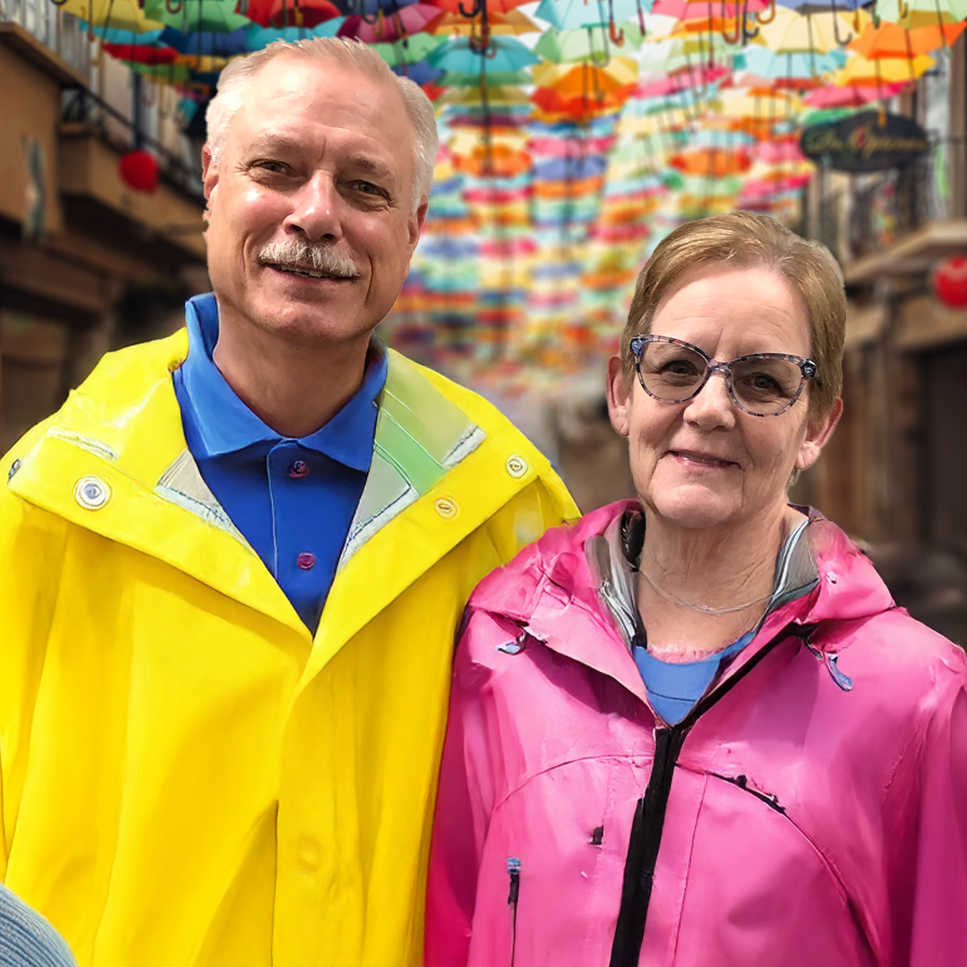 A man and woman standing together in a colorful street scene with hanging umbrellas overhead, both wearing rain jackets, the man in yellow and the woman in pink.