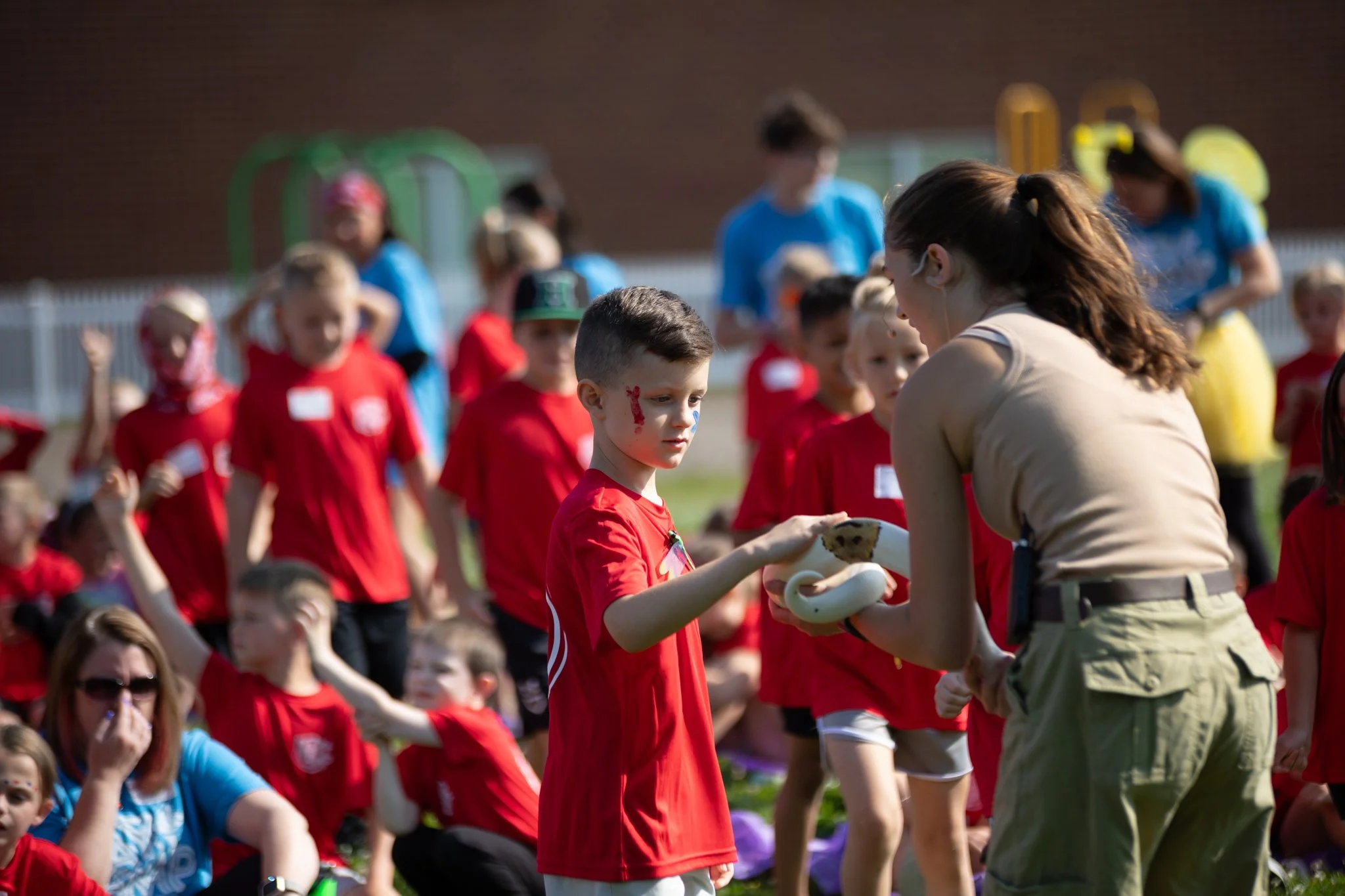 A group of children in red shirts participating in an outdoor event, with one boy receiving a white plush snake from a woman in a beige shirt. Others are sitting or standing around on a grassy field, with some in the background near playground equipm