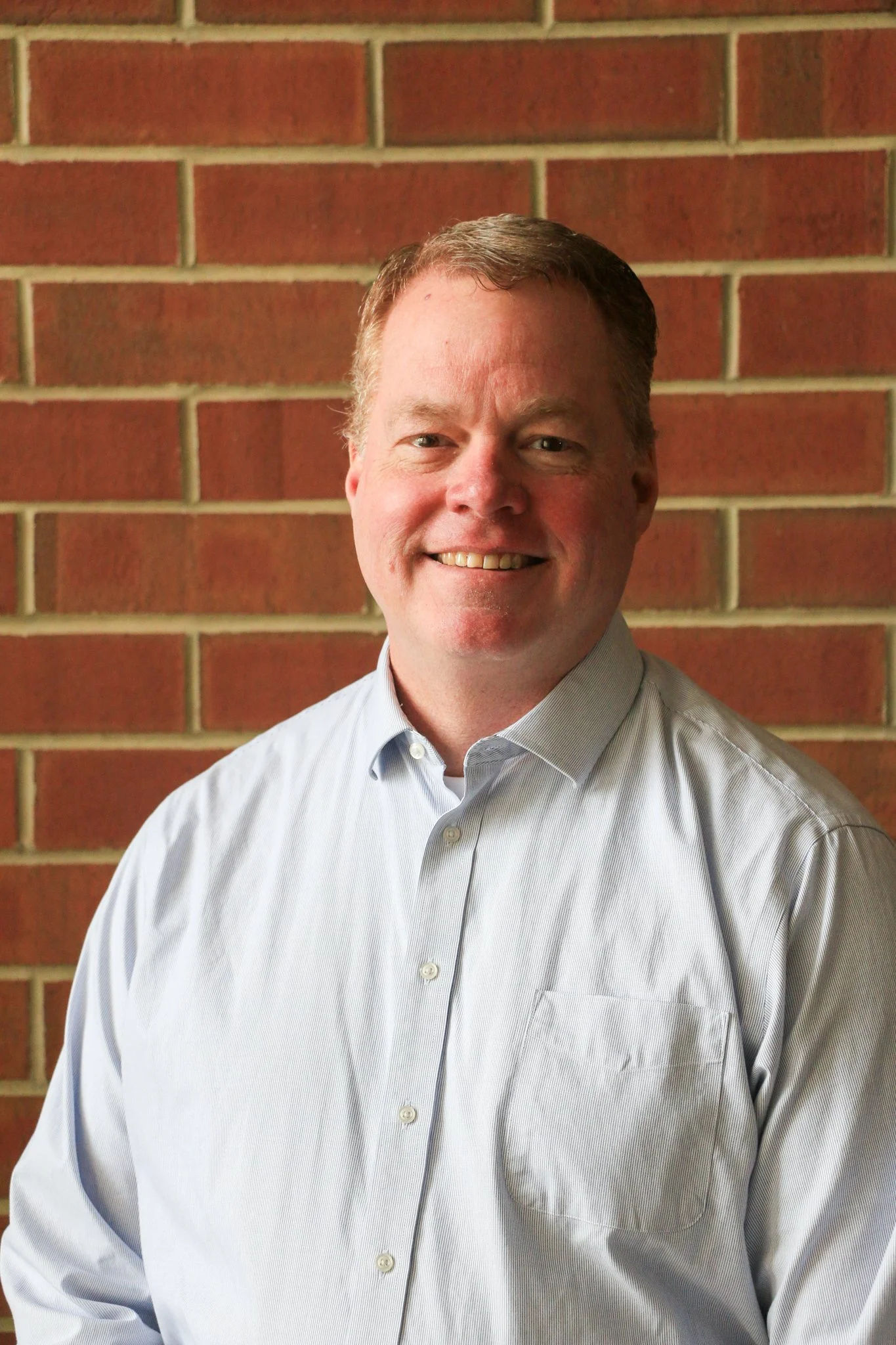 A smiling man with gray hair in a blue button-up shirt standing in front of a brick wall.