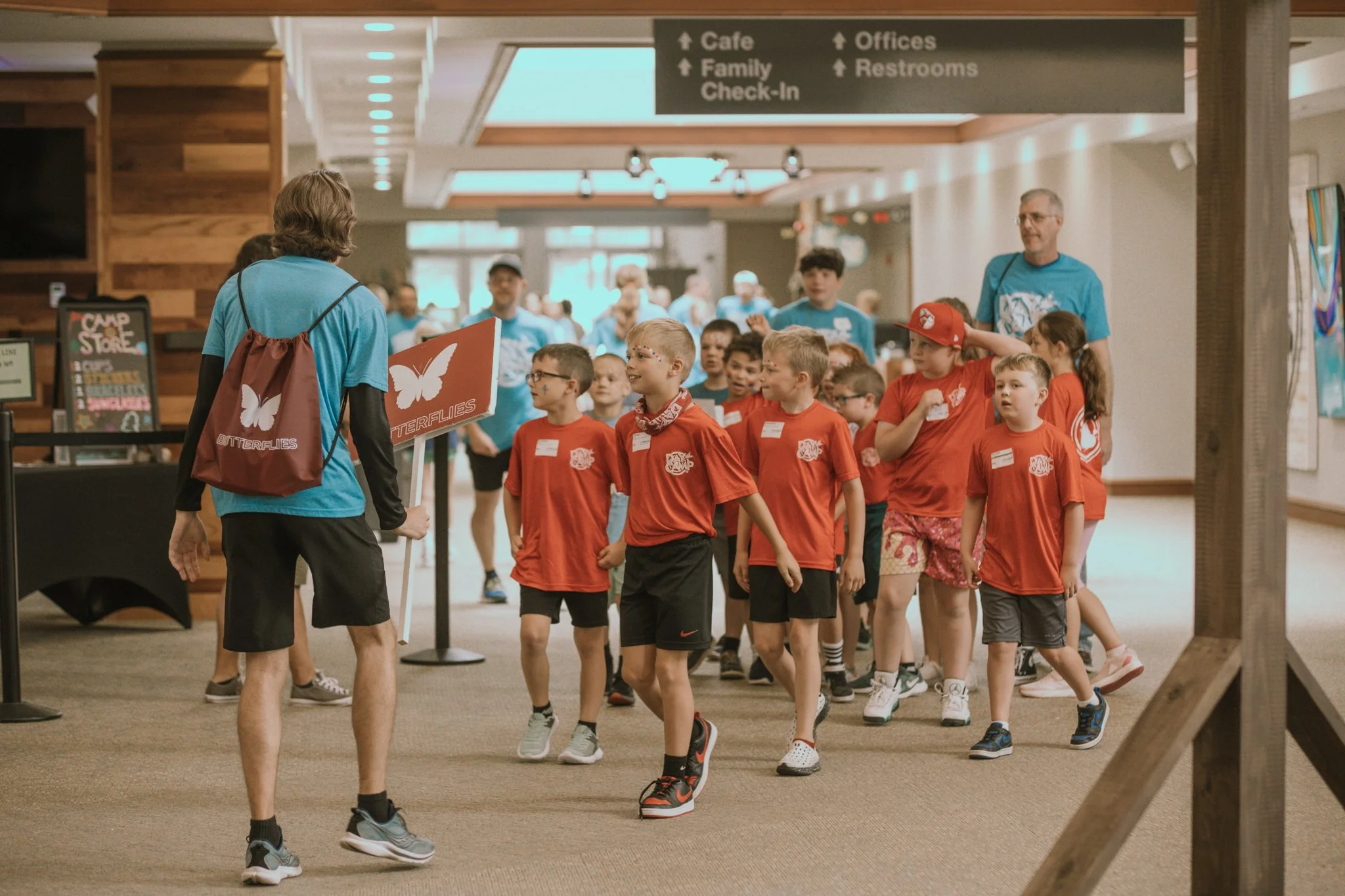 Group of children in red shirts walking in line through indoor space, guided by adult in blue shirt holding a “Butterflies” sign.