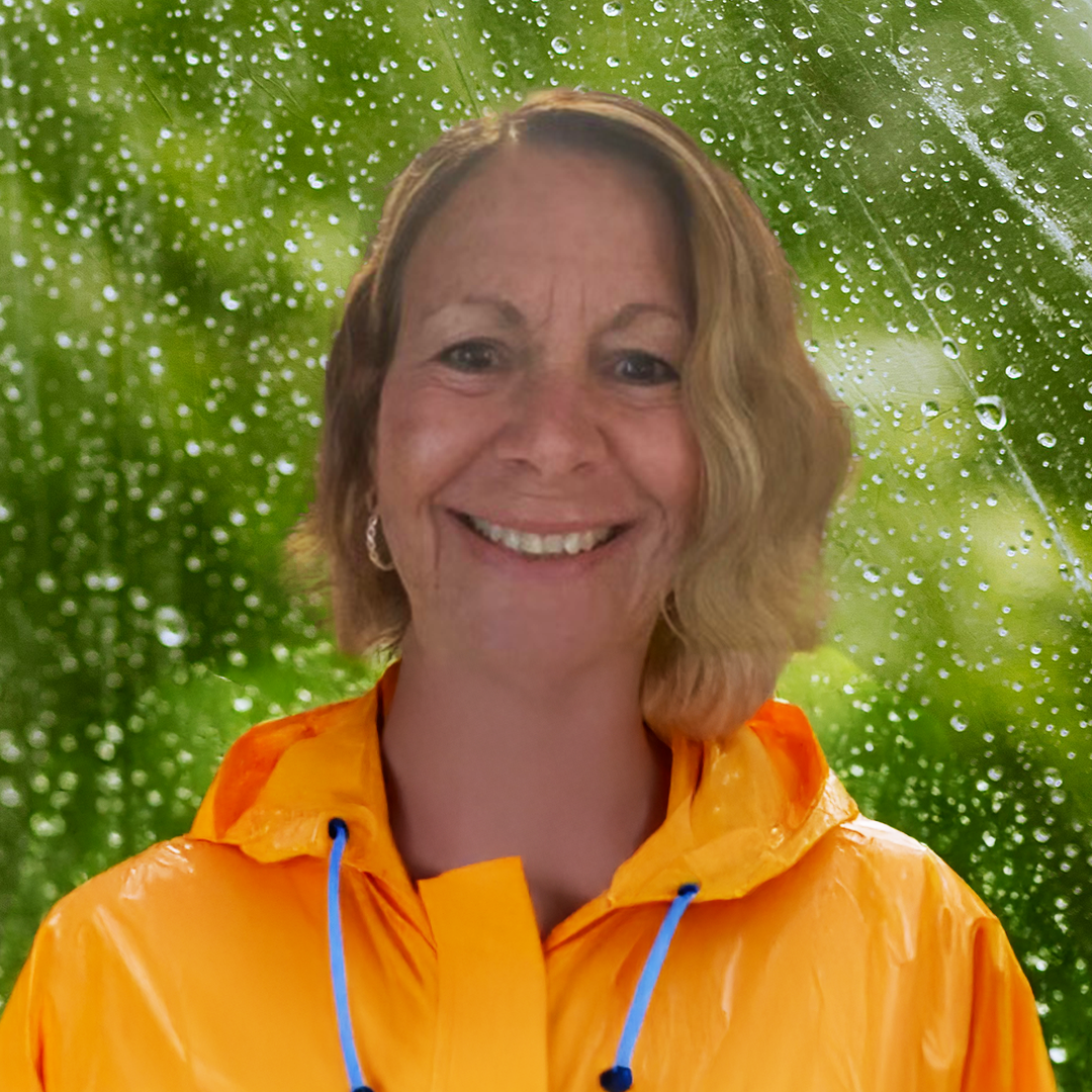 A woman with shoulder-length blonde hair smiling, wearing an orange rain jacket, standing in front of a background with raindrops on a glass surface and green foliage.