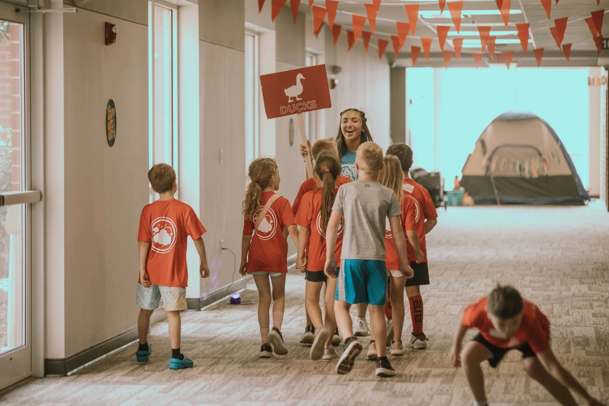 A group of children wearing red T-shirts with a cloud logo are walking in a hallway, led by a woman holding a sign that reads 'DUCKS' with a duck silhouette. Some children are holding hands, and one boy is crouched down in front.