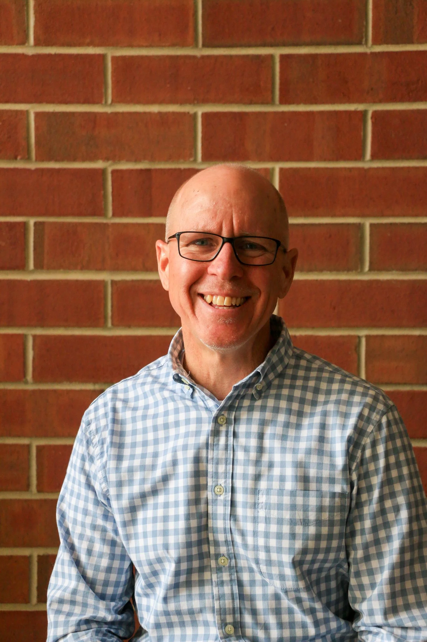 A smiling man with short hair and a beard, wearing a black shirt, standing in front of a red brick wall.