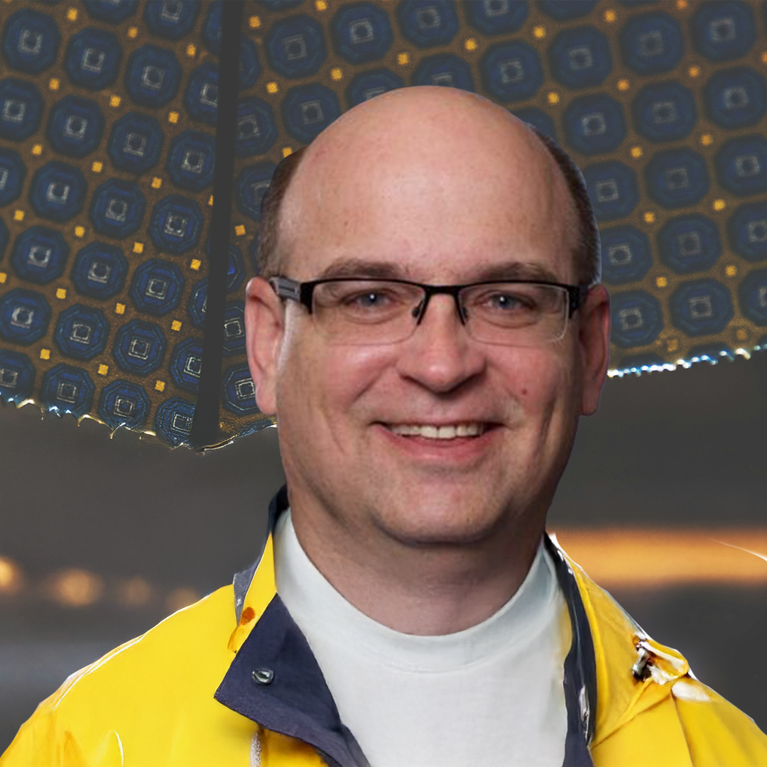 A smiling man wearing glasses, a white turtleneck, and a yellow jacket standing in front of a colorful patterned umbrella.