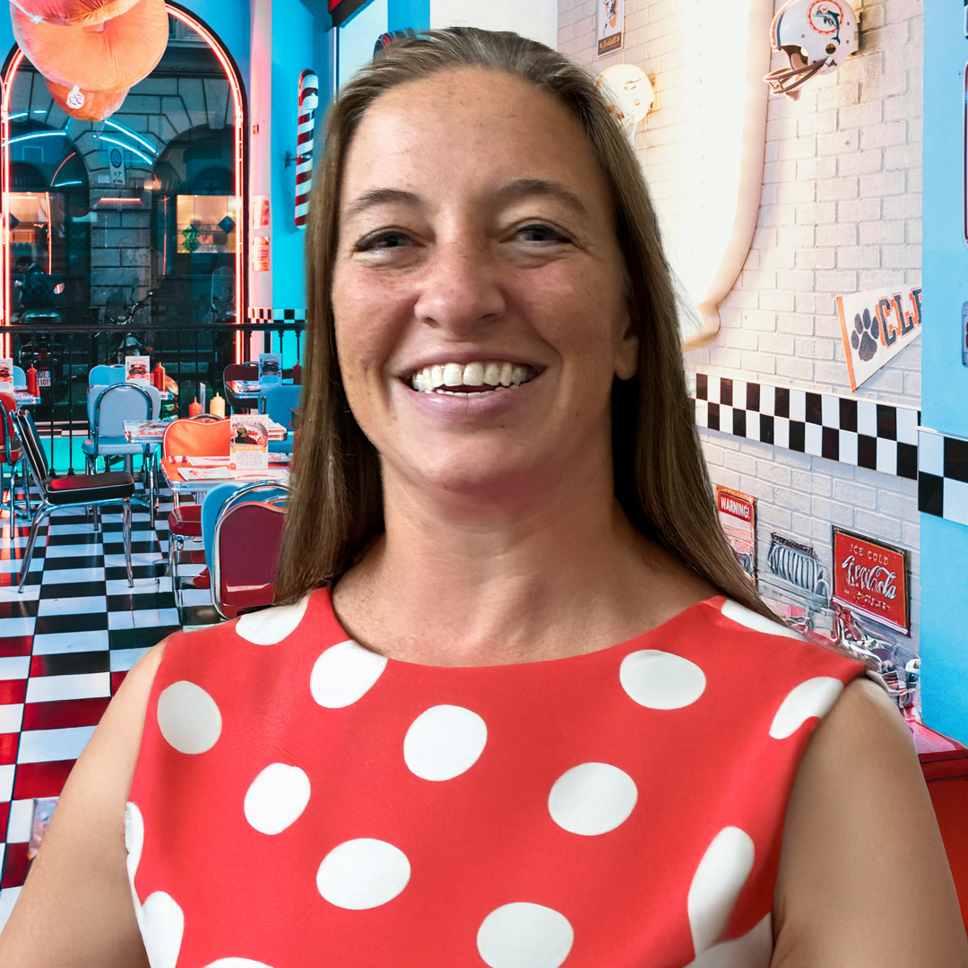 A woman with long brown hair smiling outdoors in the rain, wearing a bright green jacket over a white shirt, with a blurred green and black background.