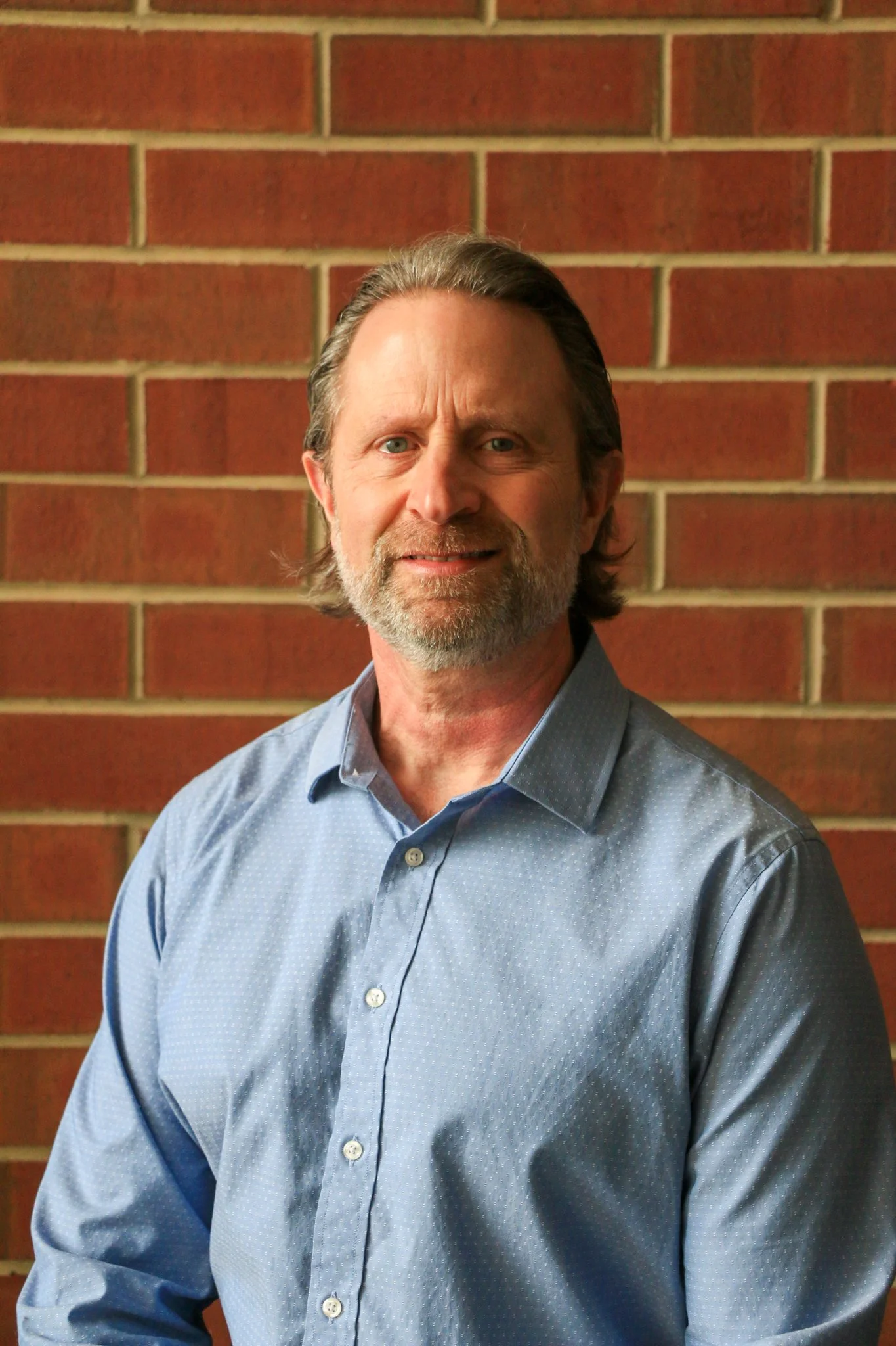A young man with short brown hair, glasses, and a black shirt smiling in front of a red brick wall.