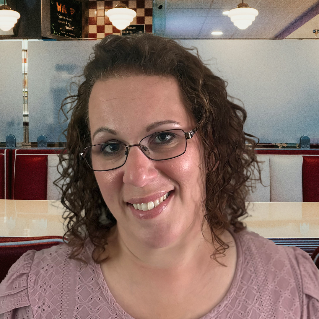 A woman with curly brown hair and glasses smiling at the camera, standing in front of a colorful background made of open umbrellas in green, blue, yellow, orange, and red.