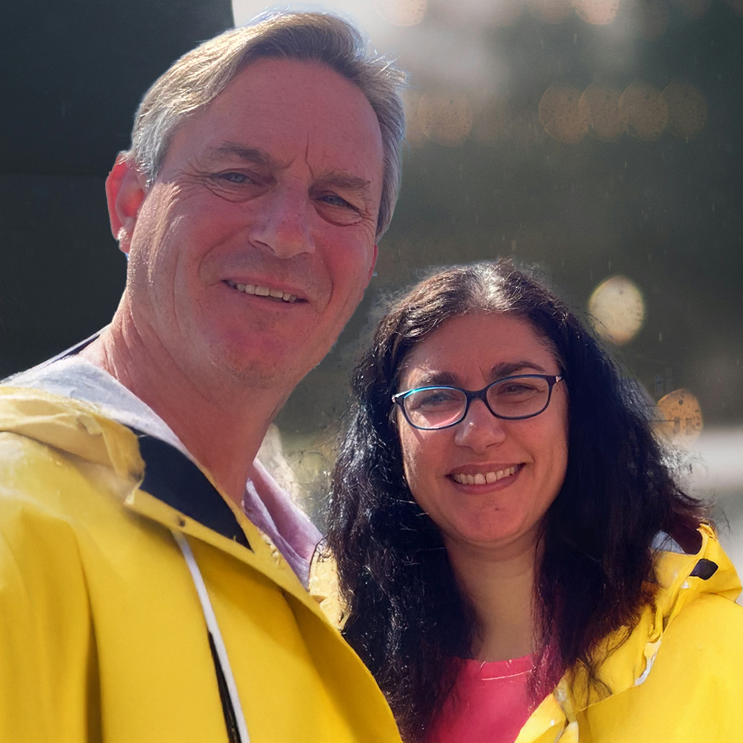 A man and woman smiling outdoors, both wearing yellow rain jackets with a blurred background and bokeh lights.