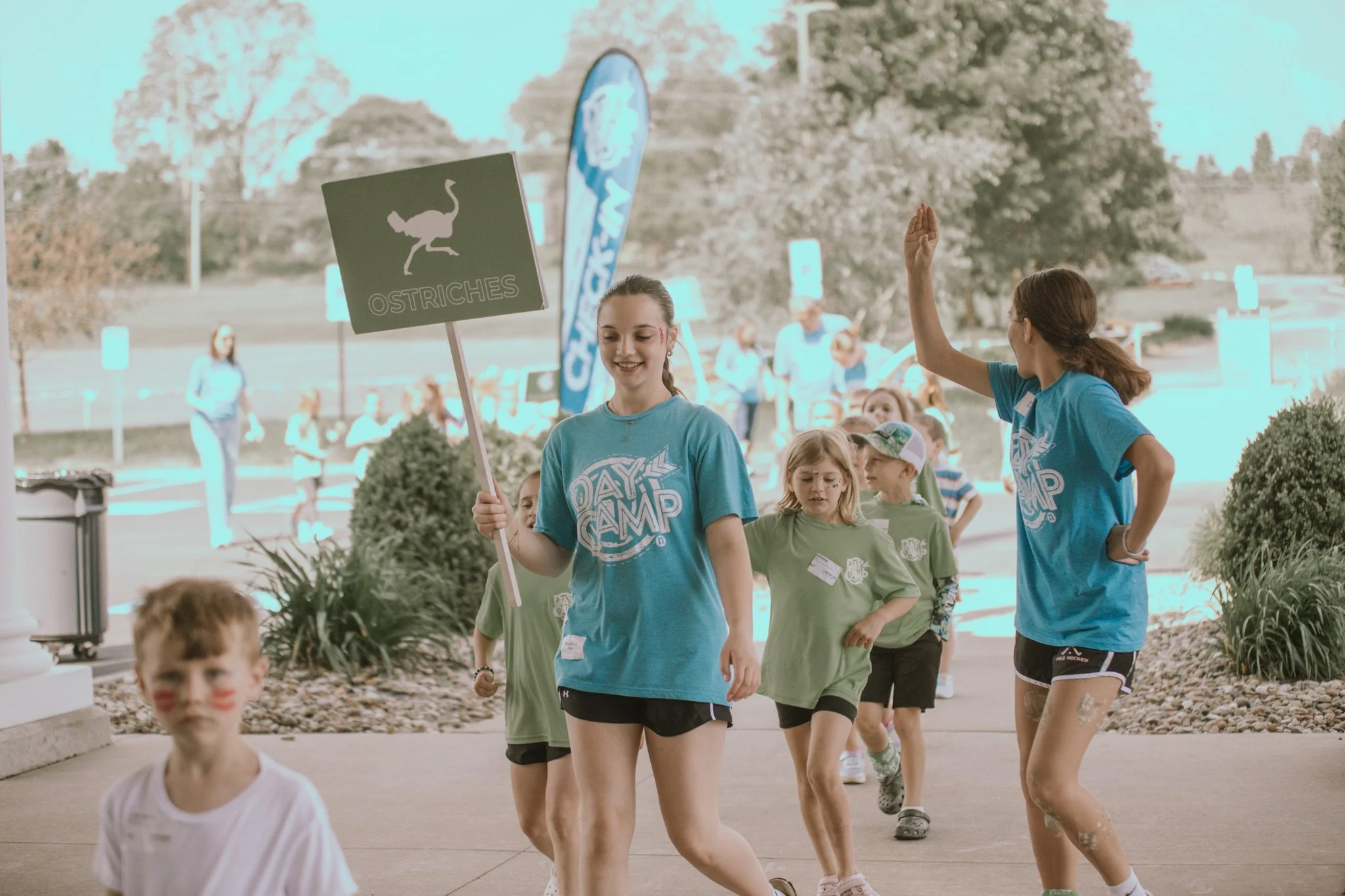 Camp counselor holding a sign labeled 'Ostriches' leading children in line outdoors at a camp event.