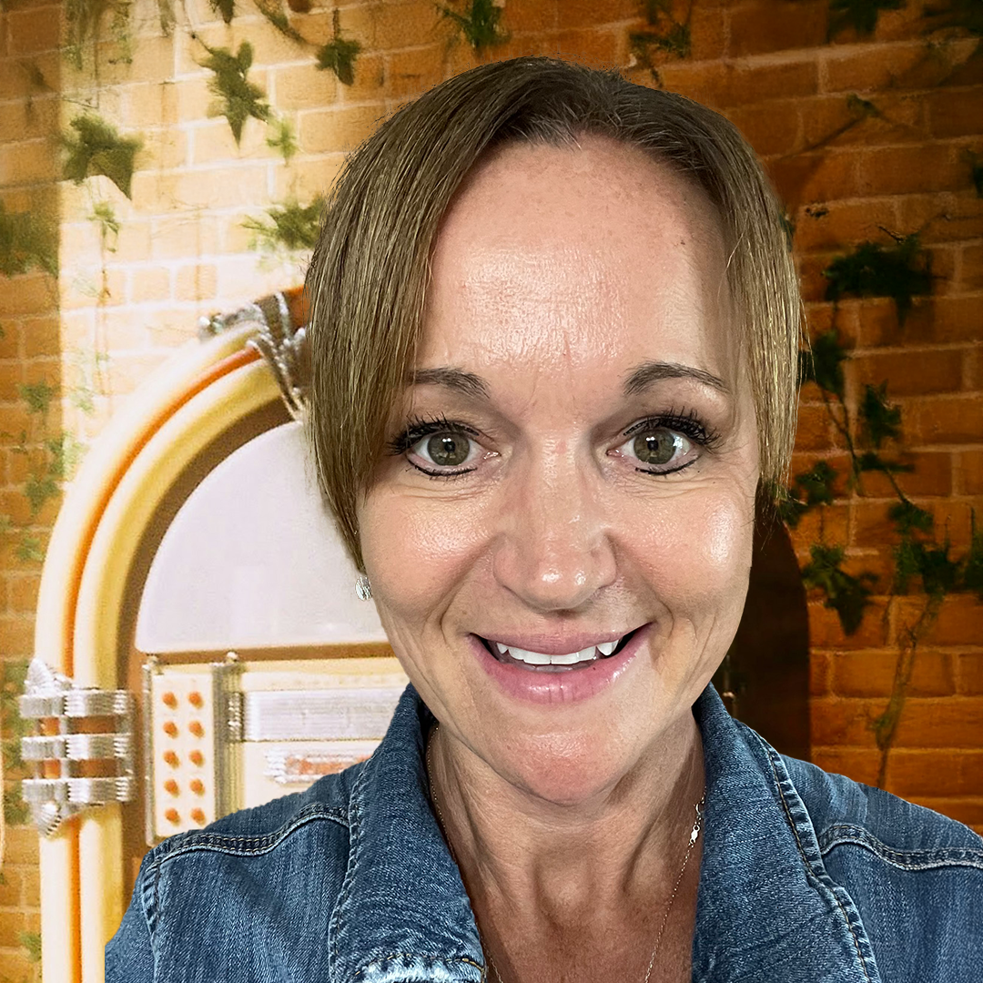 Close-up of a smiling woman with short light brown hair, wearing a bright blue jacket with yellow accents, standing in front of a window with raindrops and water streaks, with a blurred outdoor scene.