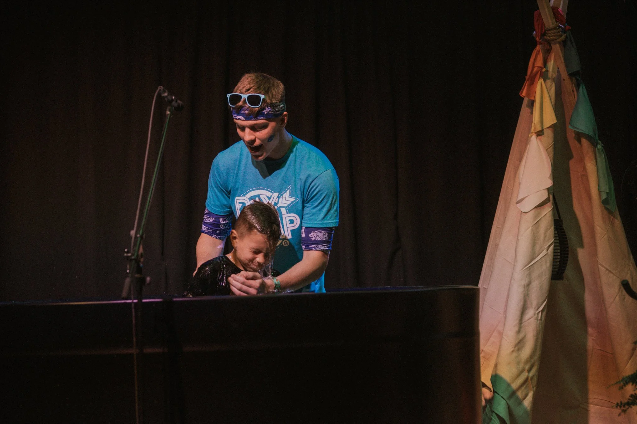 A young man wearing glasses on his head and a blue T-shirt holds a young boy over a baptismal pool during a baptism ceremony. The boy is crying tears of joy, and the young man is smiling.