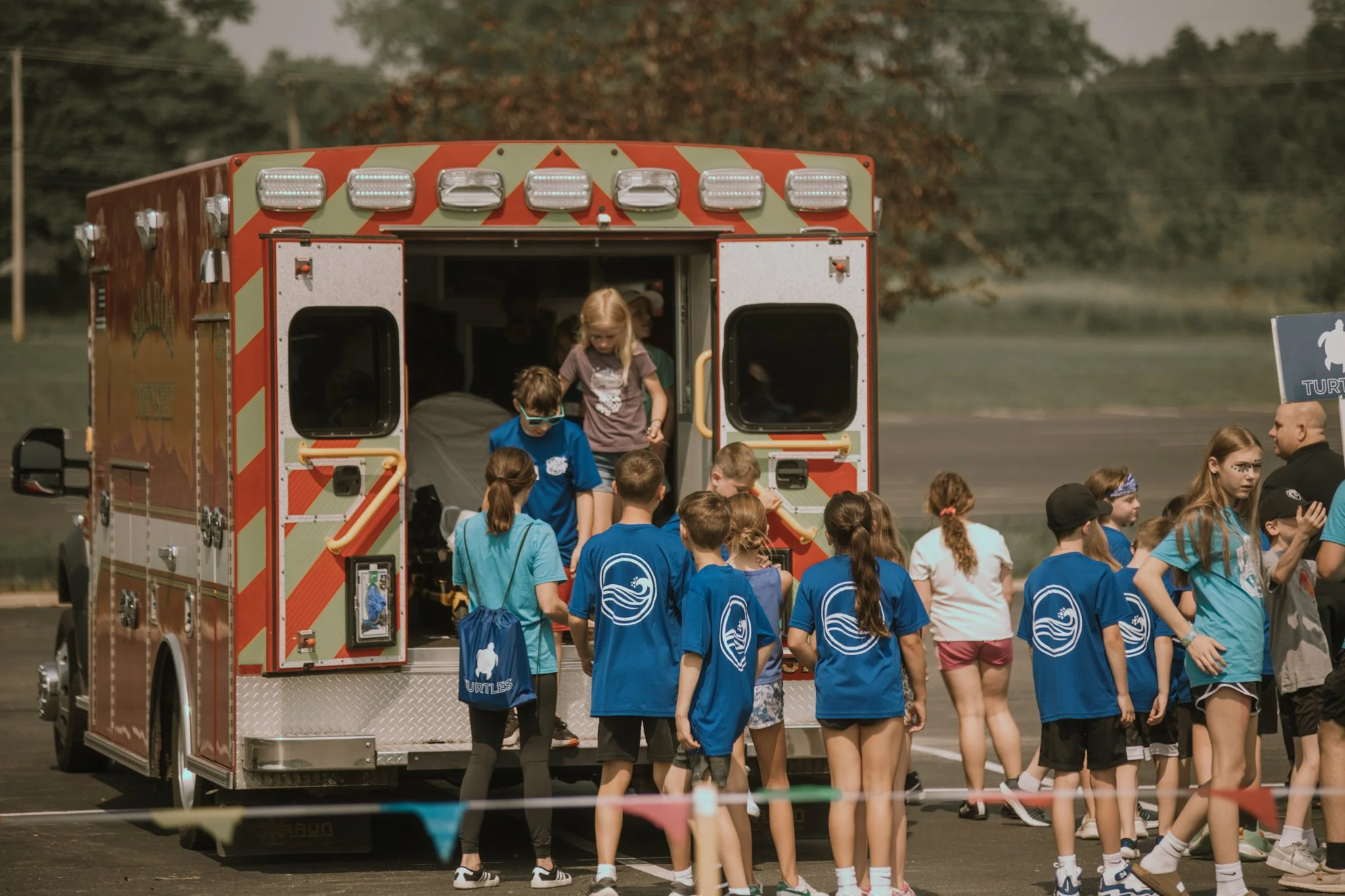 Children gather around a red ambulance on an outdoor field, some children are wearing matching blue t-shirts with a wave logo, and a girl is climbing out of the ambulance, while an adult talks to them.