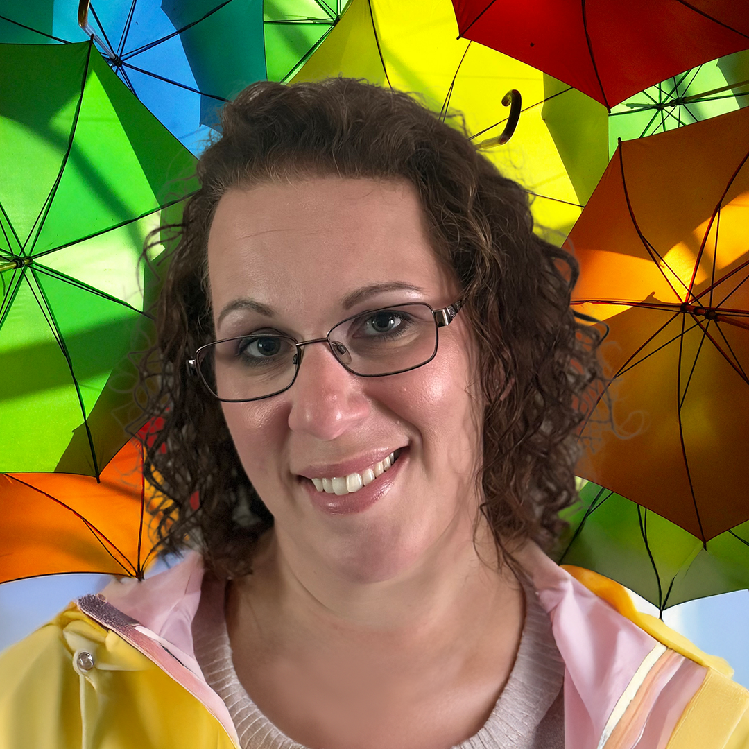 A woman with curly brown hair and glasses smiling at the camera, standing in front of a colorful background made of open umbrellas in green, blue, yellow, orange, and red.