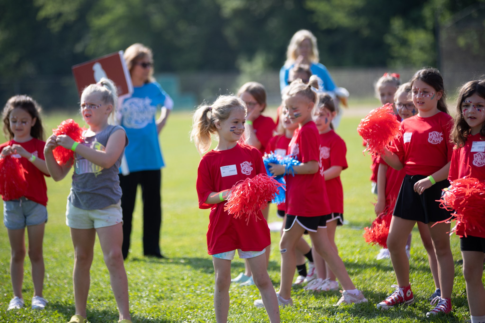 Group of young children with face paint, dressed in red shirts, holding pom-poms, standing outdoors on a grassy field with two adults in the background.