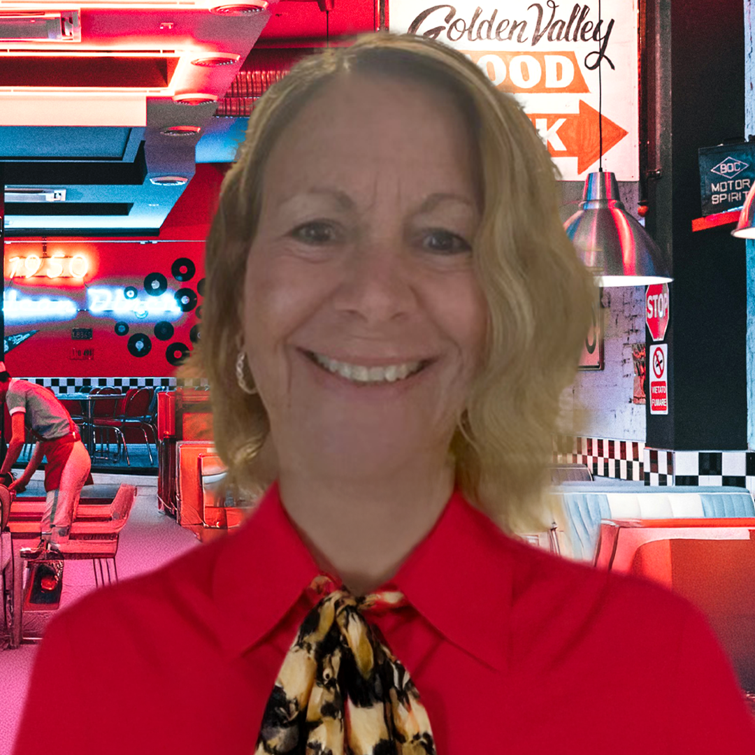 A woman with shoulder-length blonde hair smiling, wearing an orange rain jacket, standing in front of a background with raindrops on a glass surface and green foliage.