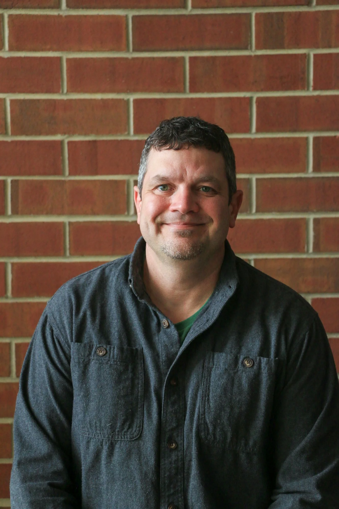 A woman with long wavy hair, wearing glasses and a black jacket, smiling and standing against a brick wall.