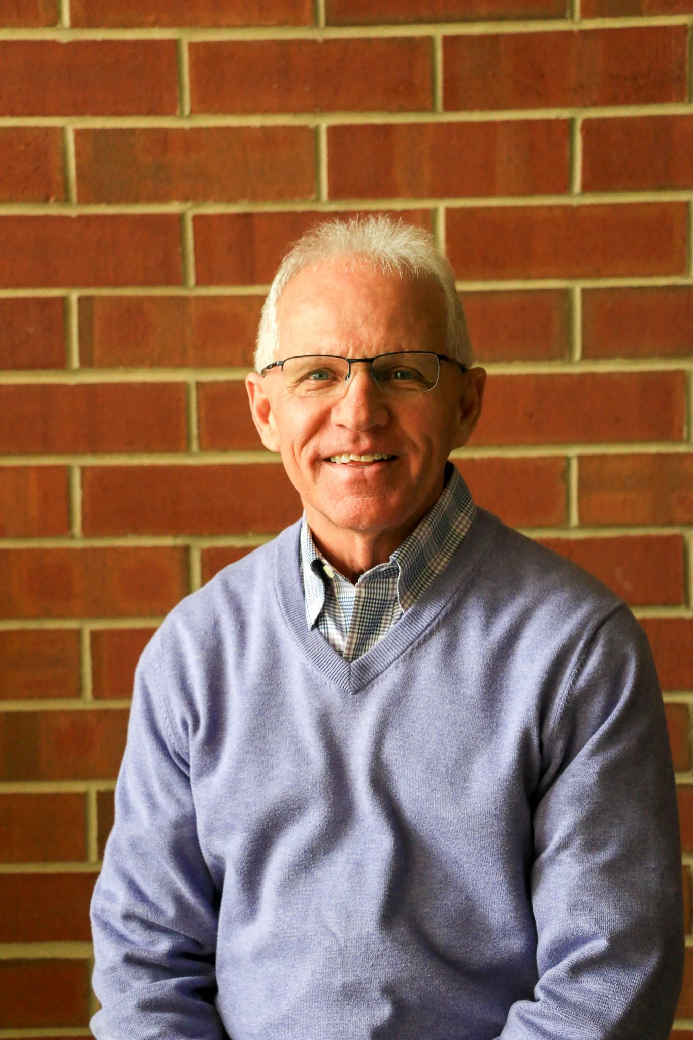 A woman with curly red hair wearing glasses and a sleeveless black top, smiling and standing against a red brick wall.