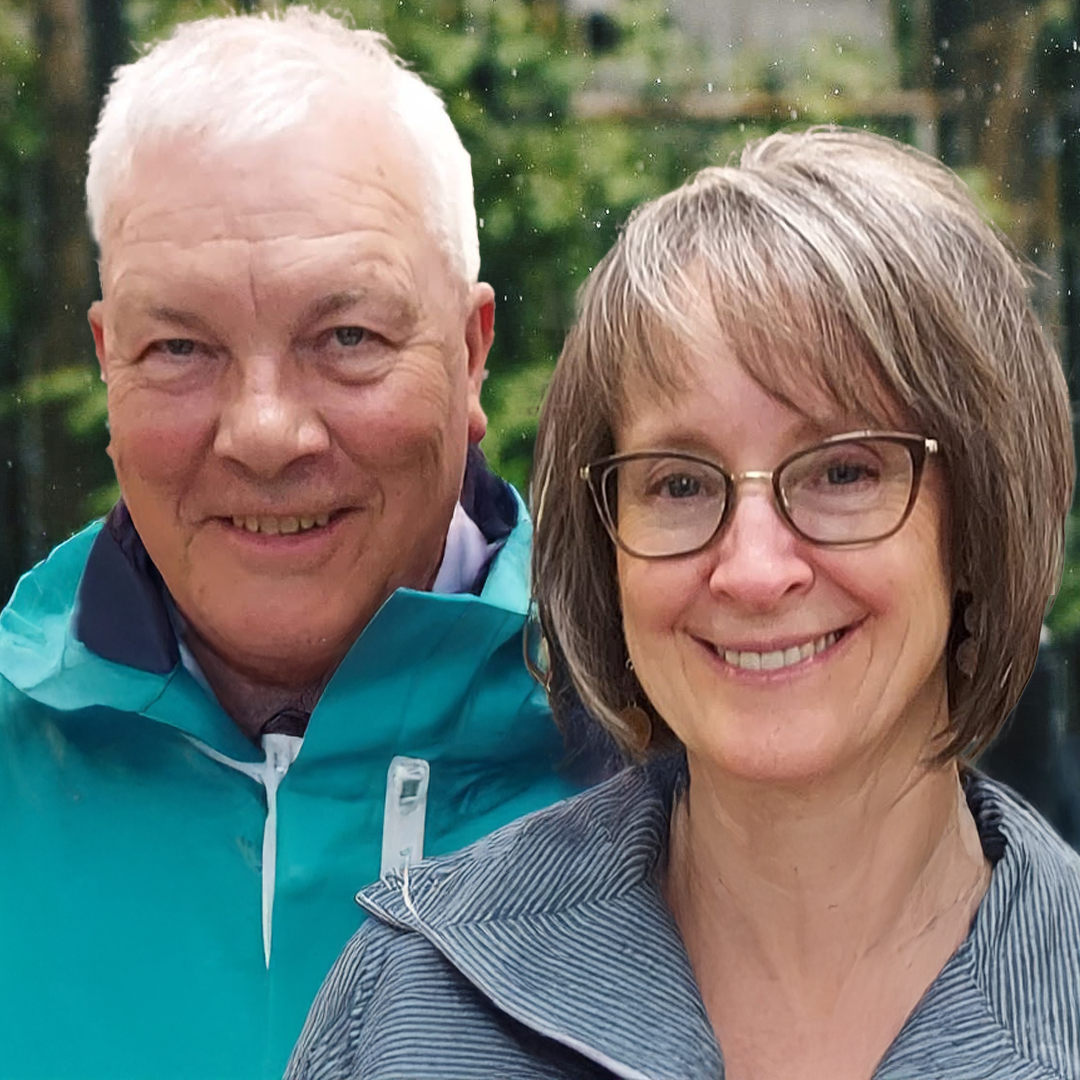 A smiling older man with white hair wearing a teal jacket, and a smiling middle-aged woman with glasses and shoulder-length hair wearing a gray symmetric striped top, outdoors with a blurred background of trees and water.