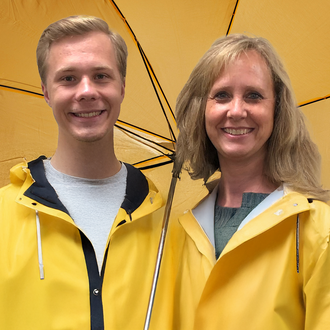 A young man and a woman are smiling while standing under a yellow umbrella. They are both wearing yellow rain jackets.