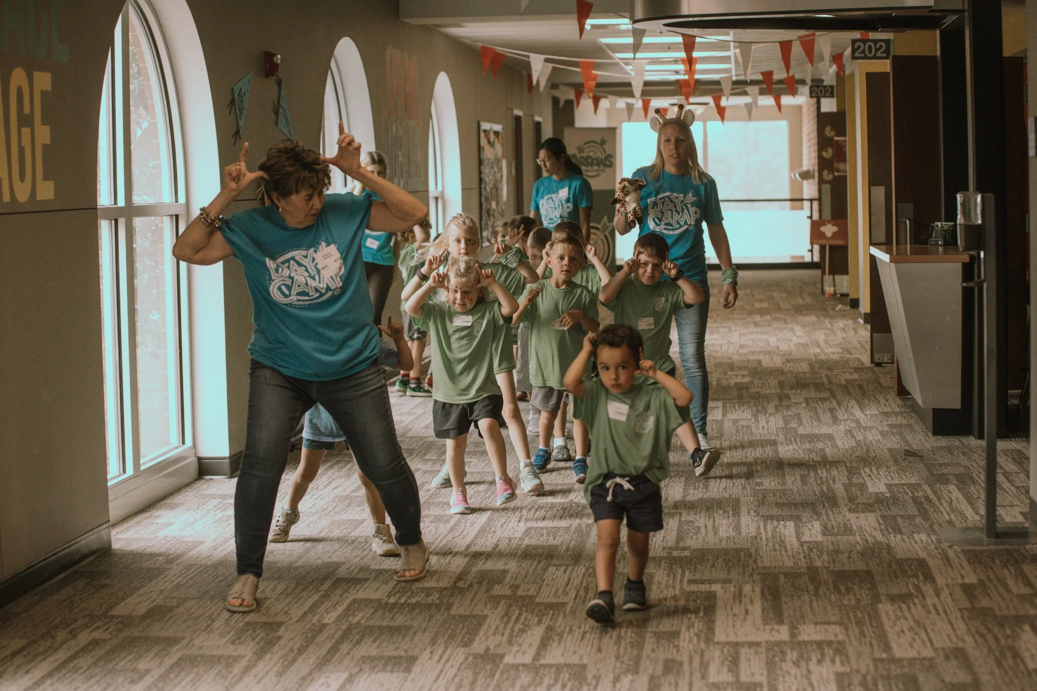 Children at summer camp walking down a hallway with two adult camp counselors leading them, some making playful gestures near their heads, in a decorated indoor setting.