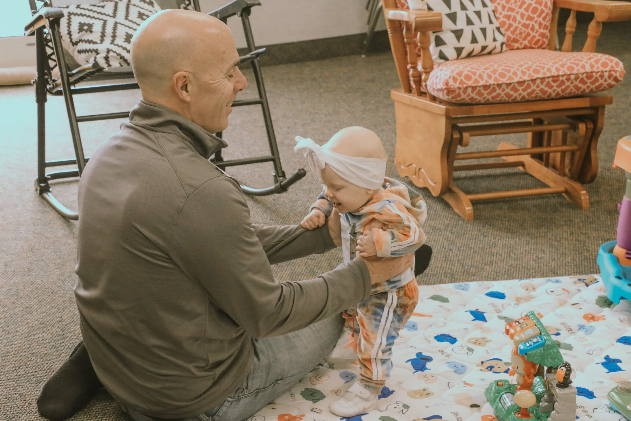 A smiling woman with glasses holding a young child in a room with a rocking chair and a playpen.
