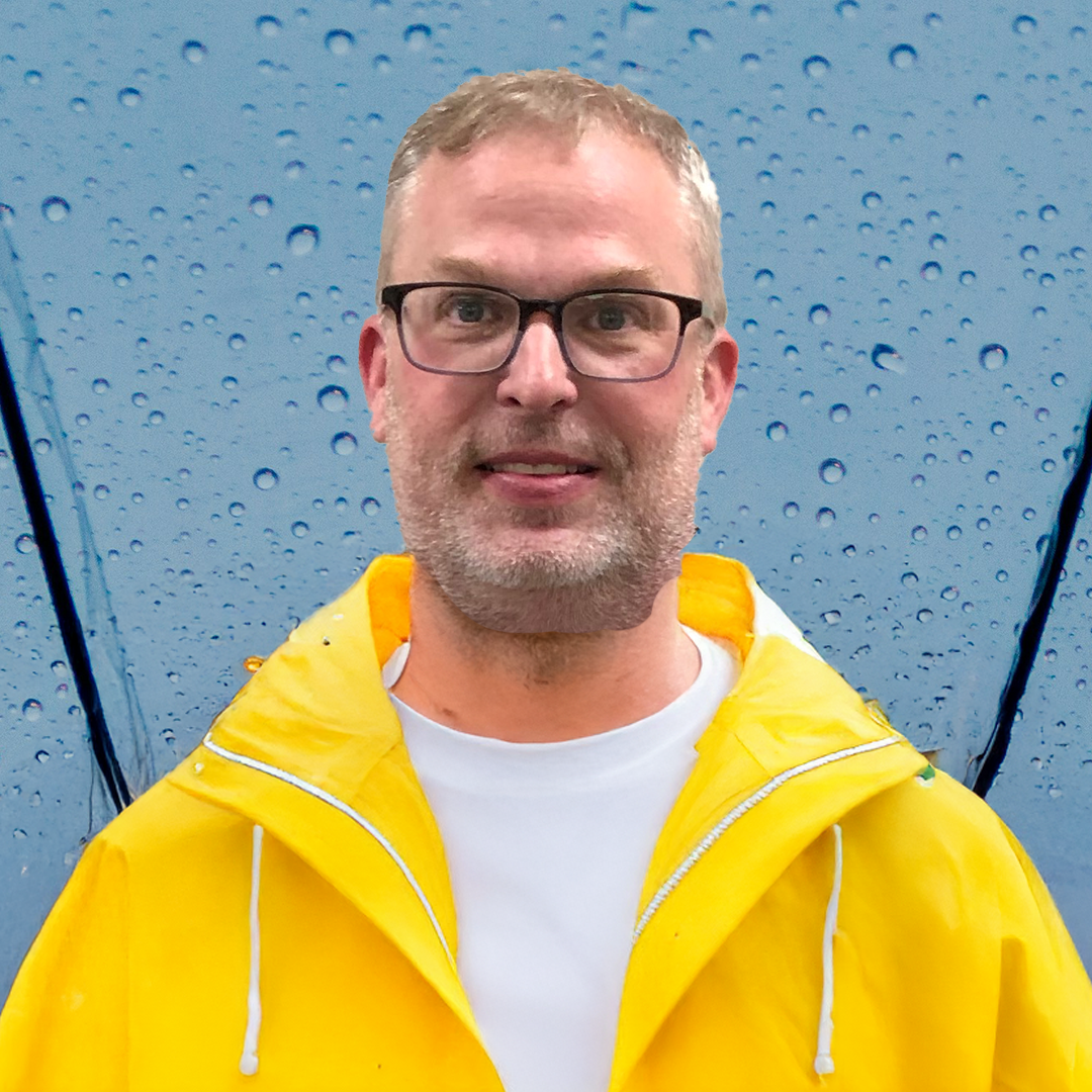A man wearing glasses and a yellow raincoat standing under a blue, rain-covered umbrella.
